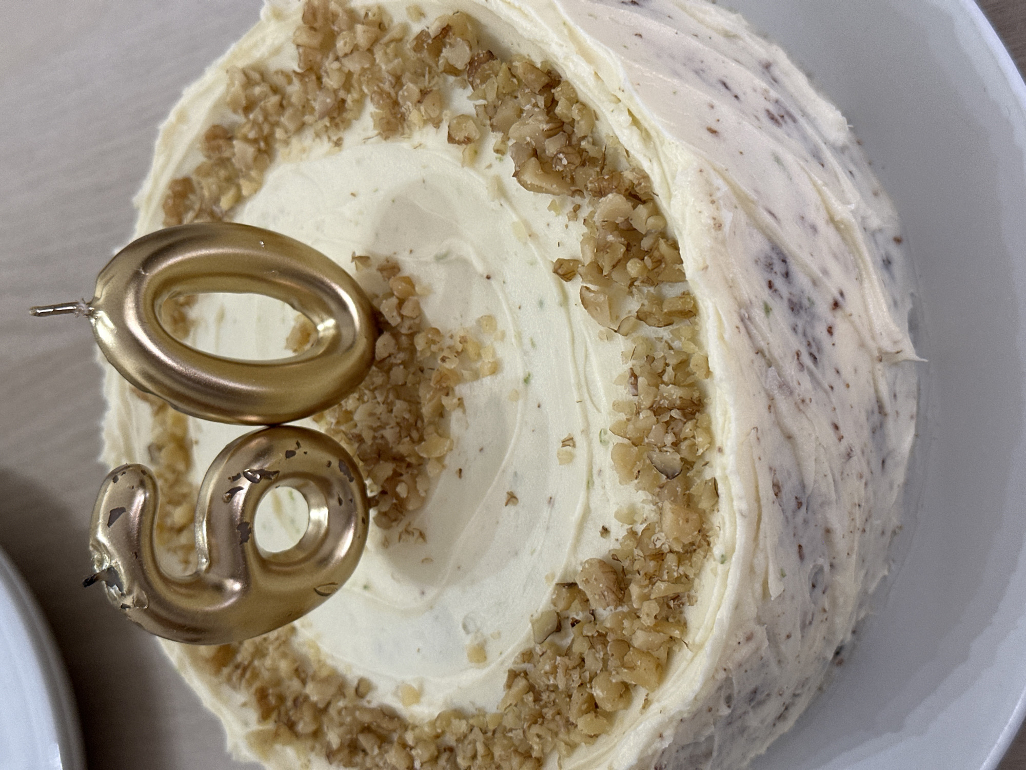 This image, captured in the city of Barcelona, Spain, depicts a celebratory cake positioned centrally on a white round plate. The cake features a generous layer of white frosting, likely cream cheese, with a subtle swirl pattern on its top surface. Sprinkled liberally over the top and along the edge of the upper surface are numerous pieces of chopped walnuts or pecans. Two gold-colored numeral candles, forming "60," are inserted into the center of the cake's top. The "6" candle shows visible wear and flaking of its gold coating, particularly around the wick and edges. The "0" candle appears slightly less worn, with a prominent wick holder. The side of the cake is also covered in the white frosting, exhibiting a slightly textured appearance with some darker specks visible through the frosting, suggesting elements within the cake layers or the frosting itself. A blurred background indicates an indoor setting, with glimpses of other white tableware. There are no people visible in the frame, and no specific actions are occurring beyond the static presentation of the cake.