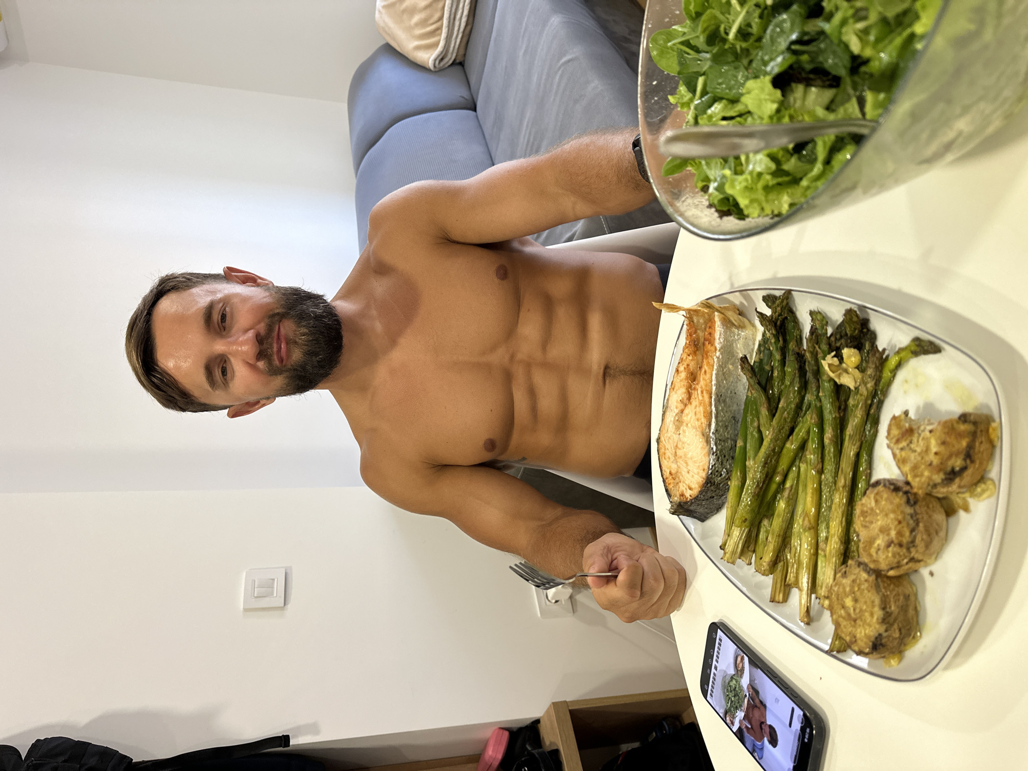 A shirtless man with a beard is sitting at a table in Donji Pobori, Montenegro, in front of a meal. The meal consists of a piece of salmon, roasted asparagus, what appear to be stuffed mushrooms or patties, and a bowl of salad. A smartphone is also on the table, displaying an image of a man holding a salad. The man in the image is holding a fork in his right hand, seemingly about to eat. In the background, a grey sofa and a white wall are visible.