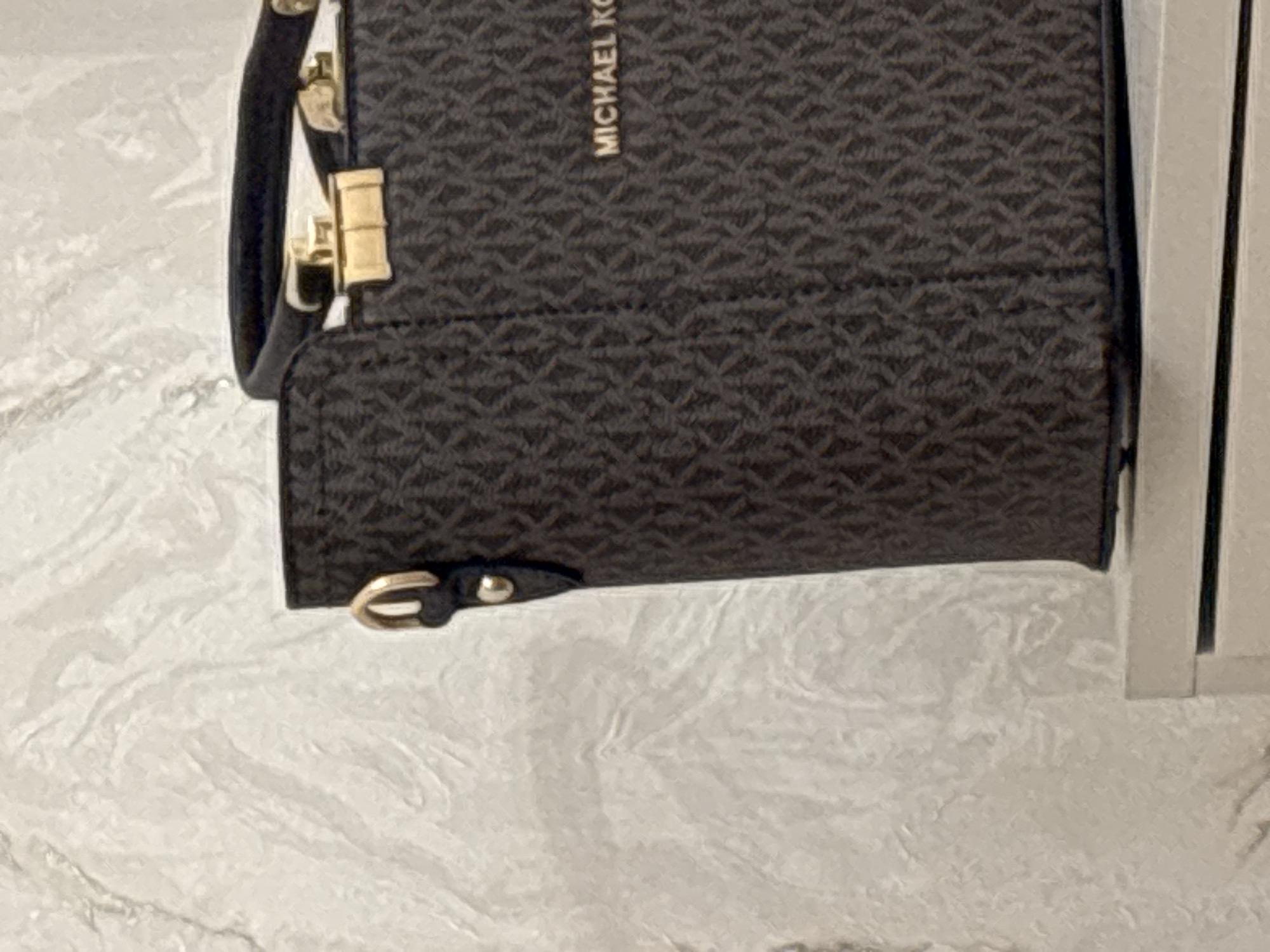 The image shows a close-up of a Michael Kors handbag with a dark brown monogram print and gold hardware. The handbag is placed on a white surface. The background is a textured white wall. The handbag has a top handle and a gold clasp. The Michael Kors logo is visible on the side of the bag in gold lettering. There is no indication of the city of Getahovit, Armenia in the image.