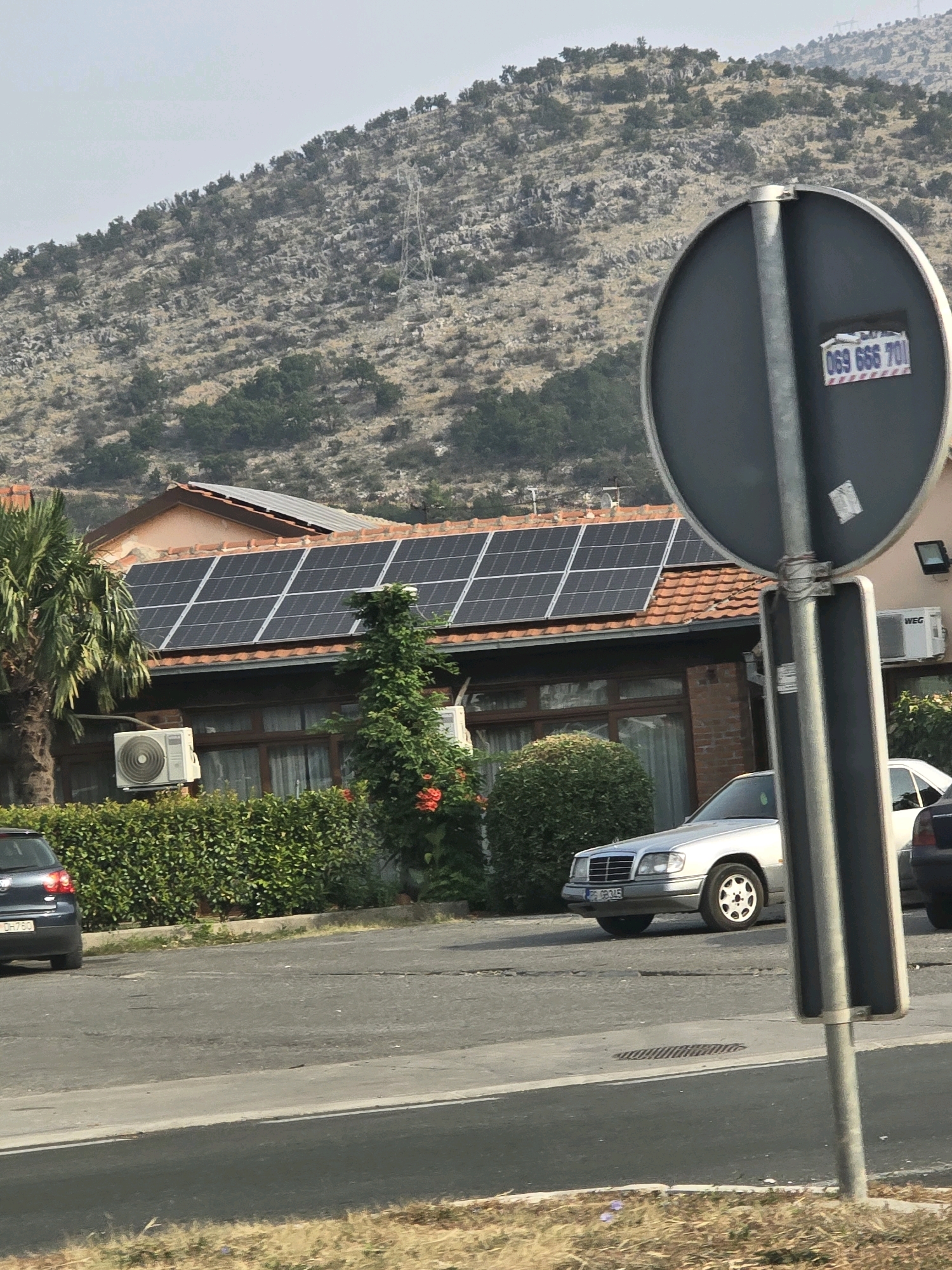 The image captures a street scene with a residential building in Podgorica, Montenegro, featuring solar panels on its roof. In the foreground, a grey car is parked on the right, and a dark car is visible on the left. A road runs in front of the building, bordered by a neatly trimmed hedge. Behind the building, a steep, rocky hillside is covered with sparse vegetation. A traffic signpost with a circular black sign is prominently positioned in the right half of the frame. The sky is overcast.