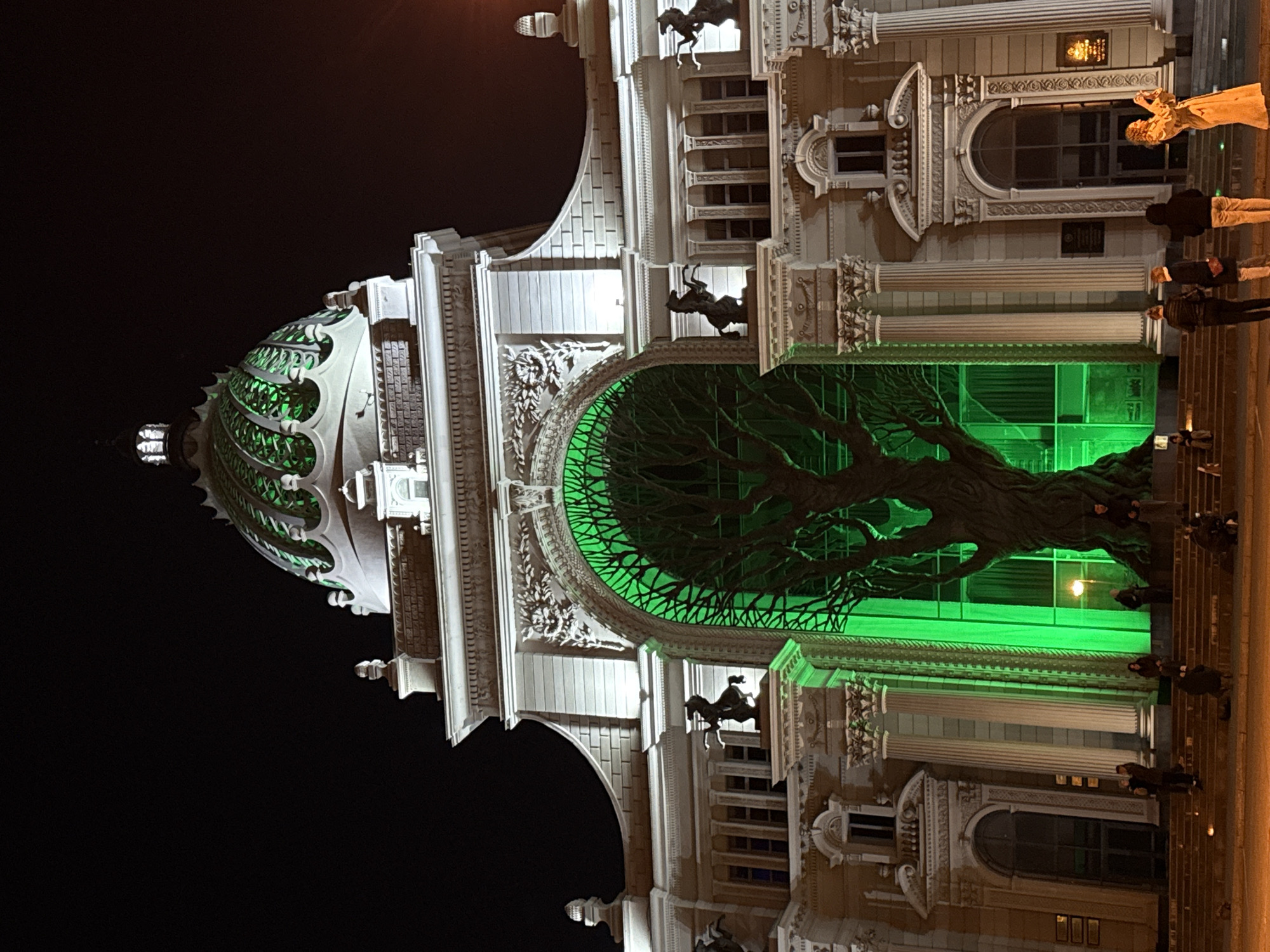 The image shows the Palace of Farmers in Kazan, Russia, illuminated at night. The building's architecture is elaborate, with a large dome on the left and multiple levels of detailed facades. A prominent feature is the metal tree sculpture within the central arch, lit with green light. Several people are visible on the right side of the frame, standing on steps and walking along the sidewalk. The sky is dark.