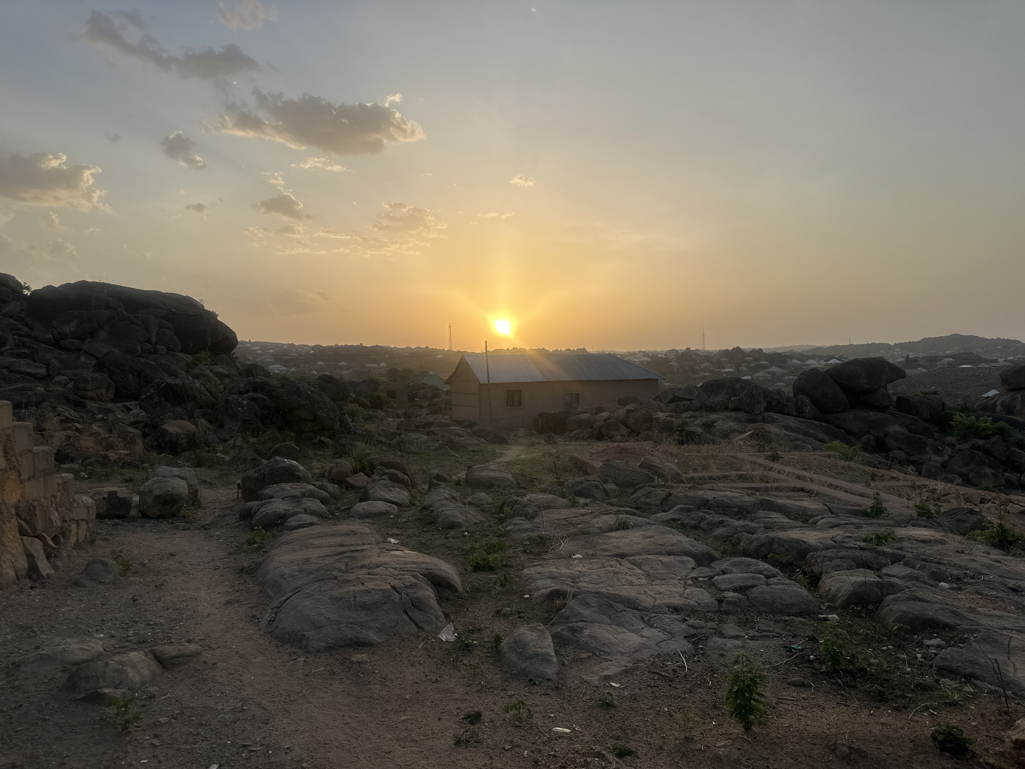 The image captures a landscape at sunset in Jos, Nigeria. The sun is positioned directly behind a small building with a metal roof, creating a bright focal point. The surrounding terrain is rocky and uneven, with scattered vegetation. In the distance, other buildings and structures suggest an urban area. The sky is a mix of yellow and light blue hues, with some clouds present. A dirt path is visible in the foreground. The overall impression is of a rural or peri-urban setting on the outskirts of Jos.