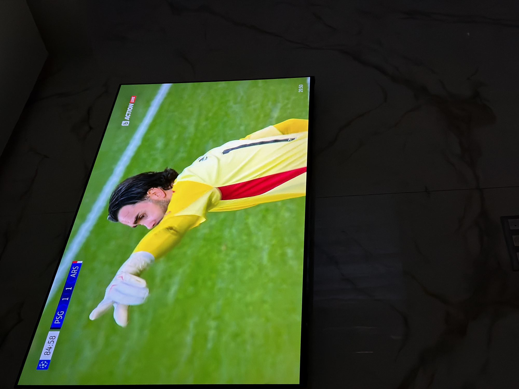 A flat-screen television displays a live sports broadcast, likely soccer, with the score at 1-1 between PSG and ARS at the 84:58 mark. The Action Live logo is visible in the upper left corner of the screen, along with the time 20:50 in the upper right corner. The scene features a goalkeeper in a yellow jersey with a red stripe, gesturing with his gloved hand. The background appears to be a green soccer field. The television is set against a background of marble-like flooring or wall. There is no indication this image was taken in Asenovgrad, Bulgaria.