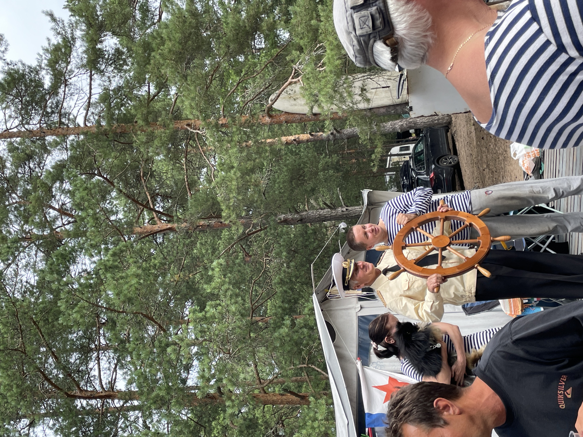 The image depicts an outdoor scene with a group of people gathered near a tent amidst a backdrop of tall pine trees. A man in a naval-style uniform, complete with a white hat and gold trim on his jacket, stands holding a wooden ship's wheel. Beside him, another person in a striped shirt and gray pants smiles. A woman holding a small, fluffy black dog stands to the left of the man in uniform. In the foreground, a person wearing a black t-shirt with a logo is partially visible. In the background, a dark-colored car is parked, and a white trailer or RV is also present. The overall setting suggests a casual gathering or event in a wooded area, possibly a campsite in Zelenaya Roshcha, Russia, given the presence of pine trees and the mention of the location.