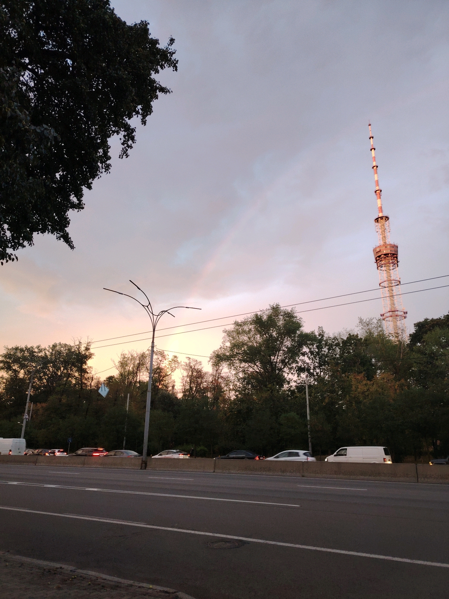 An overcast sky with a faint rainbow arcs over a broad, multi-lane road in Kyiv, Ukraine. A large tree dominates the upper left of the frame, its dark foliage silhouetted against the pale sky. To the right, a tall television tower, painted with white and red bands, pierces the sky, its intricate lattice structure visible. A line of cars and vans travels along the road, their headlights and taillights reflecting the muted light. Dense trees line the far side of the road, forming a verdant backdrop. A streetlamp with multiple curved arms stands prominently near the center, its design minimalistic and modern. The overall impression is one of an urban landscape at dusk, with a touch of natural beauty in the form of the rainbow.