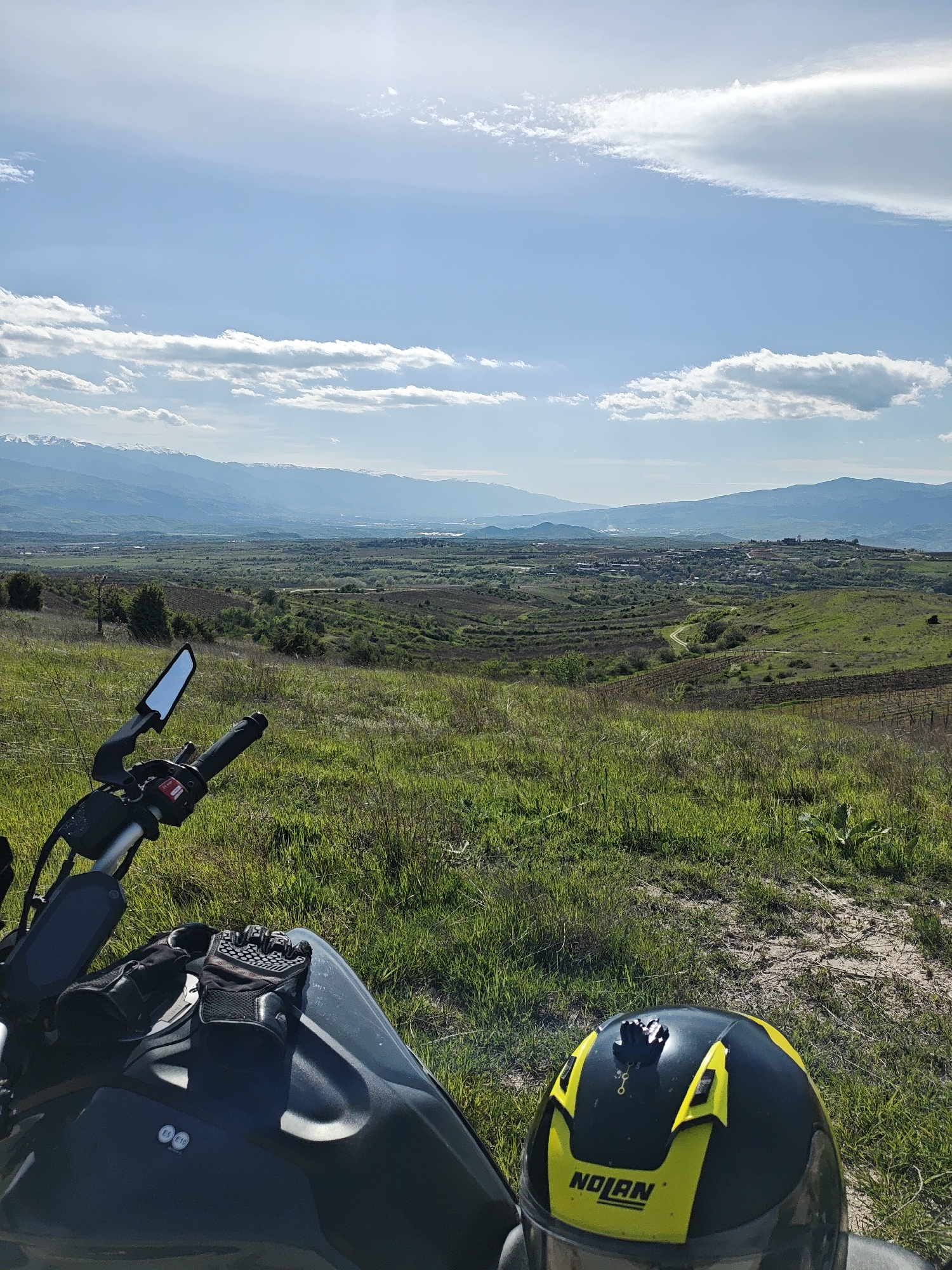 The image captures a scenic view from what appears to be a grassy hillside. In the foreground, a portion of a motorcycle is visible, including the handlebars, a mirror, and a pair of gloves resting on the tank. A Nolan brand motorcycle helmet, predominantly black with yellow accents, sits on the ground near the motorcycle.
The midground consists of a verdant landscape, featuring rolling hills and fields dotted with patches of trees and what appears to be vineyard rows. A dirt path meanders through the landscape.
In the background, there is a vista of mountains with some snow-capped peaks visible in the distance. Above the mountains, the sky is blue with scattered white clouds. The image suggests a rural or semi-rural environment. There is no other visible location information.