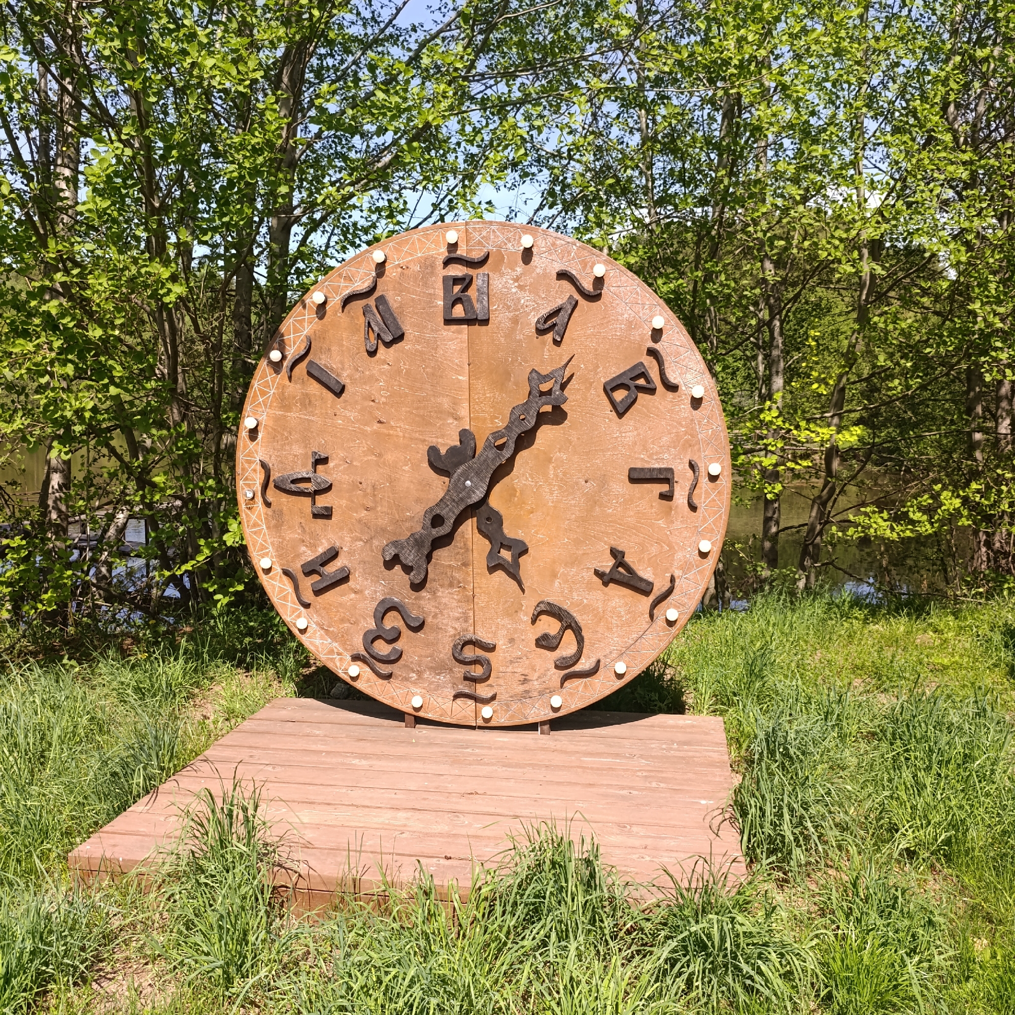 A large, round, brown clock stands on a wooden platform surrounded by green grass in Prikamskii, Russia. The clock's numbers are represented by Cyrillic letters in dark brown, and its hands are made of dark metal with decorative cutouts. The clock is positioned in front of trees, with a glimpse of a body of water visible through the trees. The scene is well-lit, suggesting it is daytime.