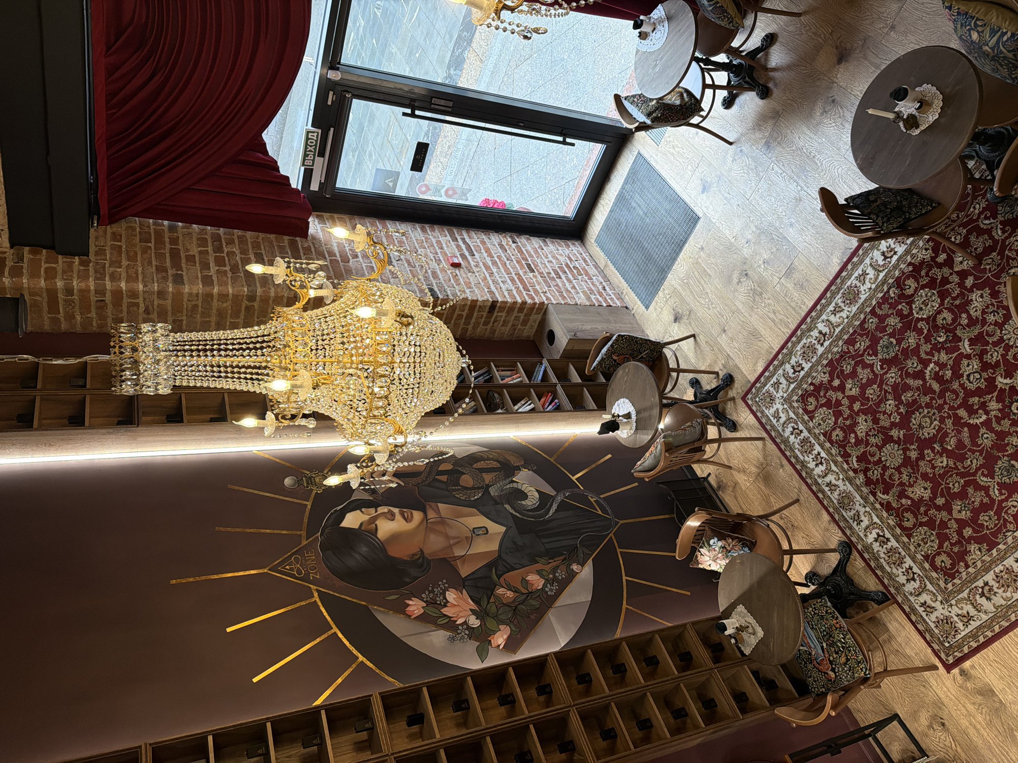 The image depicts the interior of a cafe or restaurant in Moscow, Russia. The scene is taken from an elevated perspective, looking down into the space. A prominent feature is a large, ornate crystal chandelier hanging from the ceiling. The walls are decorated with a mural depicting a woman and a snake, surrounded by geometric patterns. Underneath the mural is a shelf unit with multiple compartments, possibly for displaying bottles. The floor is made of wooden planks and partially covered by a red patterned rug. There are small round tables with chairs arranged around the rug. A glass door leads to the outside, revealing a street scene with pedestrians and vehicles. The interior design elements suggest a sophisticated and stylish ambiance.