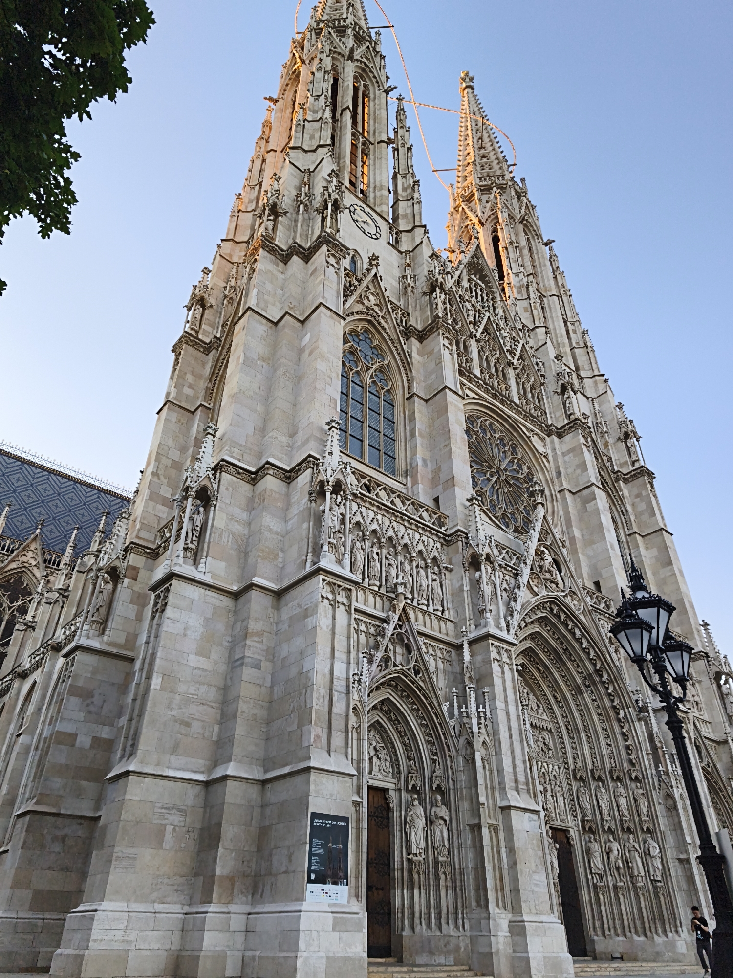 The image captures a low-angle, upward view of the Votivkirche (Votive Church) in Vienna, Austria, bathed in the warm light of what appears to be either sunrise or sunset. The church, a prominent Gothic Revival landmark, dominates the frame with its intricate stone facade, spires, and ornate carvings. The building's pale stone is illuminated, highlighting the detailed sculptures and architectural elements. A large rose window is visible on the facade, along with multiple smaller arched windows. The roof of a section of the church, with its dark, patterned tiles, is visible on the left. A tree with green leaves partially obstructs the upper left corner. In the foreground, a black street lamp with two globes stands to the right of the church entrance. Two wooden doors are set within deep arched portals. A sign with text and images is mounted on the lower left section of the church's stone wall. A lone individual is visible in the lower right corner of the image, near the base of the street lamp, appearing to look at their phone. The sky above is a clear, pale blue, with no clouds. Decorative string lights are draped between the spires of the church.