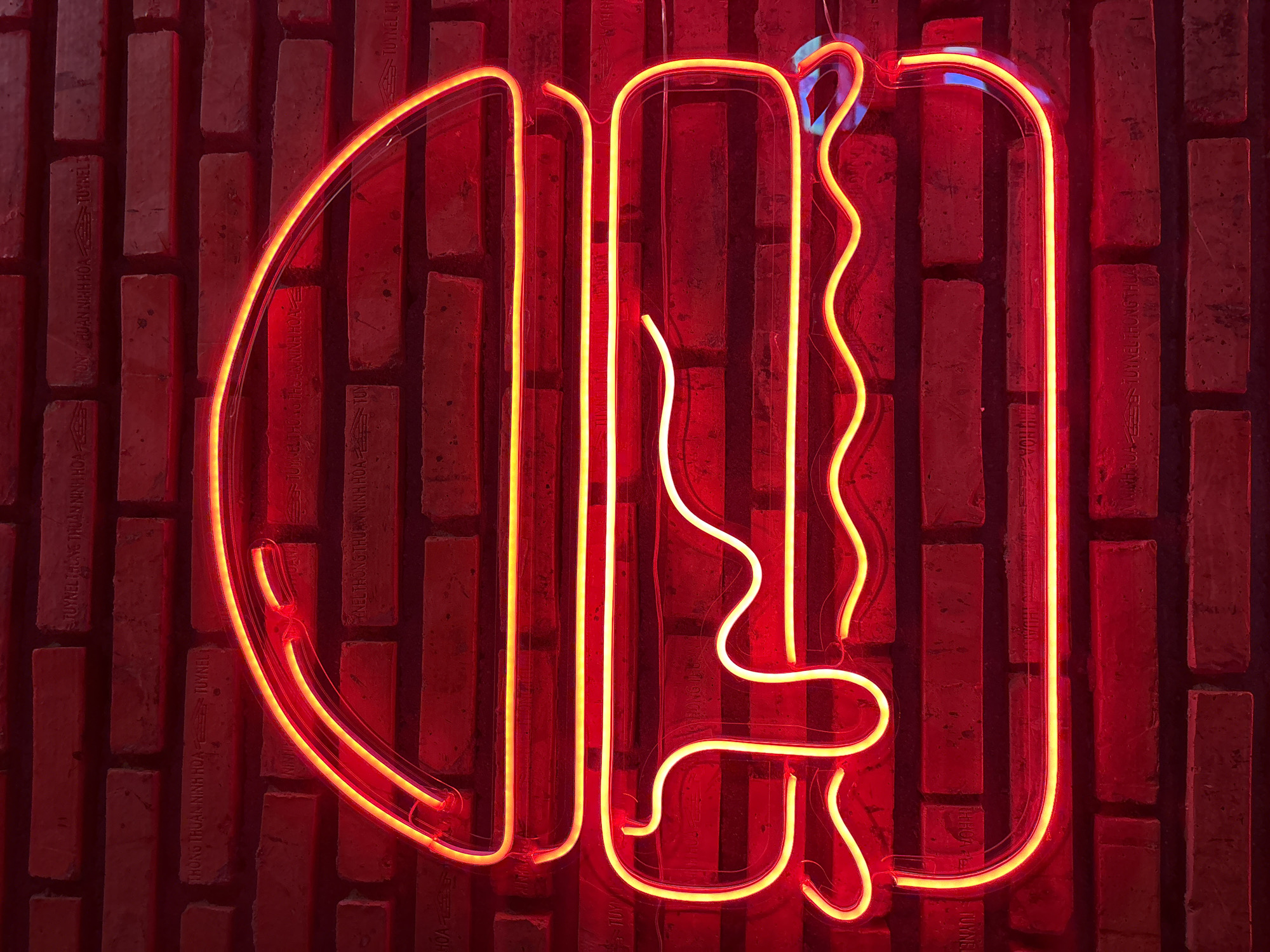 The image shows a neon sign shaped like a hamburger, glowing red and yellow against a brick wall. The sign is positioned centrally in the frame and is illuminated, casting a warm glow on the textured red brick. The hamburger design is simplified, outlined with neon tubes to represent the bun, patty, cheese, and lettuce. Some parts of the sign, like the cheese and sauce, are depicted with wavy lines and dripping effects. The brick wall behind the sign appears to be made of red bricks with visible mortar lines and some imperfections, contributing to a rustic or industrial aesthetic. The overall lighting suggests an indoor setting, likely a restaurant or bar, given the neon sign's subject matter and style. There are no people or animals visible in the image. The background consists solely of the brick wall, with no other discernible objects or elements. The image is focused on the neon sign and its immediate surroundings, highlighting the vibrant colors and design.