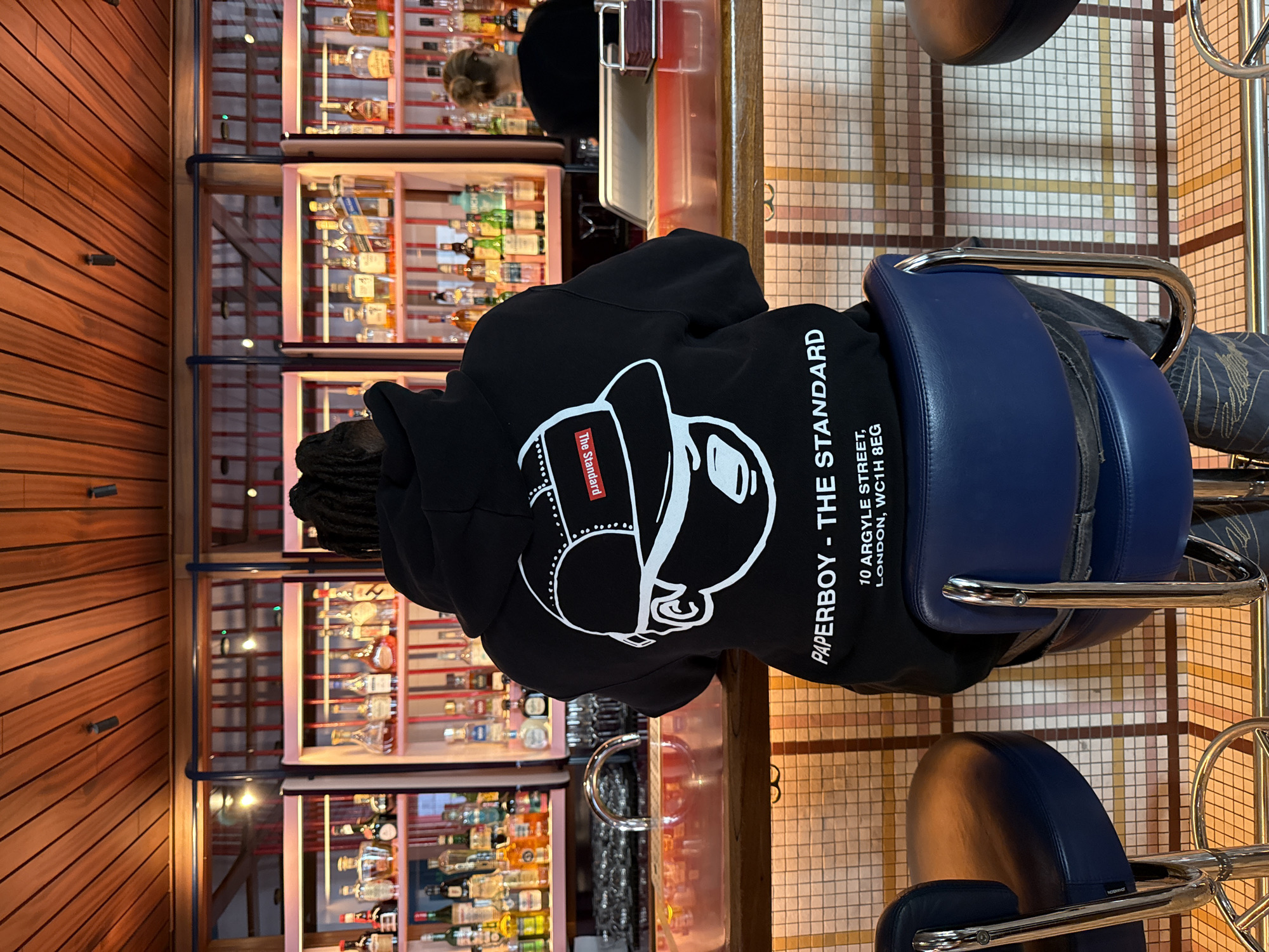 The image captures an interior scene within a bar or restaurant, presented as if the original photo was rotated 90 degrees clockwise. In the foreground, a person with dreadlocks is seated on a blue leather bar stool at a counter, facing away from the viewer. They are wearing a black hoodie featuring a large white graphic of a stylized head in a cap and glasses. Visible text on the back of the hoodie includes "PAPERBOY - THE STANDARD", along with an address: "10 ARGYLE STREET, LONDON, WC1H 8EG". A small red tag on the graphic design reads "The Standard".
Behind the seated person, a well-stocked bar is visible with multiple shelves filled with various bottles of spirits and liquors, illuminated by soft lighting. A second person, likely a bartender, is partially seen in the background behind the bar, appearing to be at work. The environment features warm-toned wooden paneling on one wall and a distinctive tiled pattern on the floor and wall behind the bar, composed of small, light-colored square tiles. The setting suggests an evening or nighttime ambiance due to the artificial lighting.