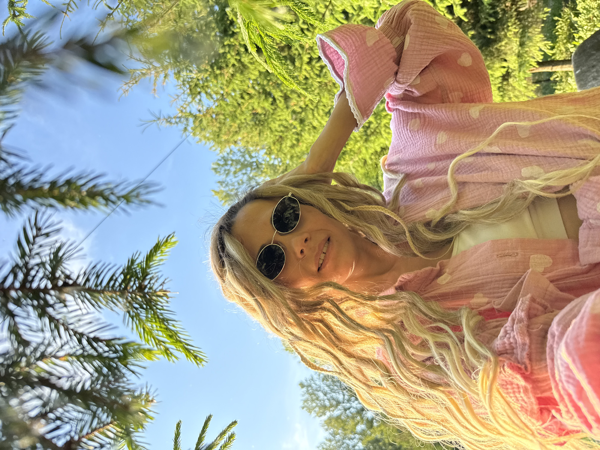 A woman with long blonde dreadlocks and sunglasses is captured in a low-angle, outdoor selfie. She is wearing a pink, polka-dot shirt and a white top. The background consists of lush green trees and a clear blue sky, suggesting a natural, park-like setting. The sun is shining, casting a warm glow on her hair. The image does not contain any information about the city of Rusanovo, Russia.