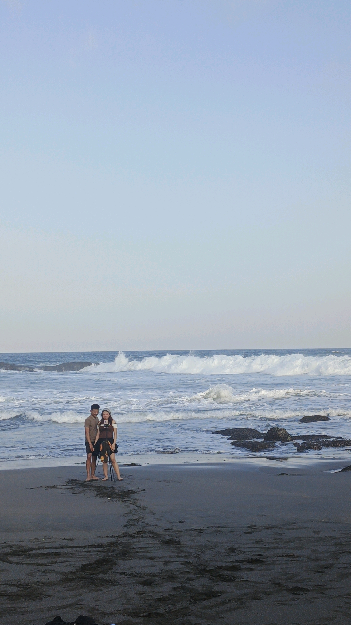 A young couple is standing on a dark sand beach with the ocean in the background. The man is wearing a brown t-shirt and shorts, and the woman is wearing a black dress and a white headscarf. The woman is holding a phone and appears to be taking a selfie. Waves are crashing on the shore, and there are some rocks visible in the water. The sky is clear and pale blue. The scene appears to be in a coastal area.
