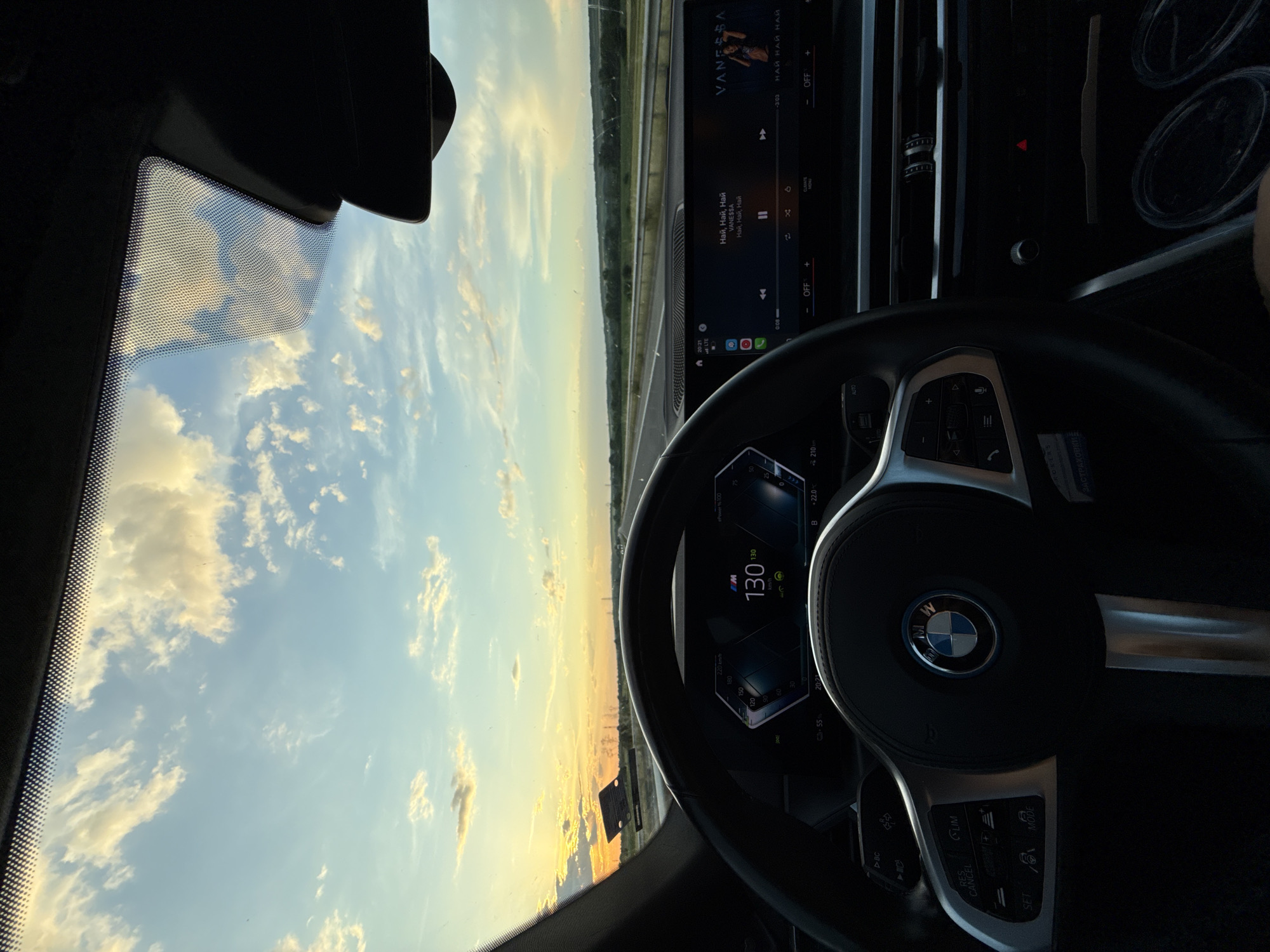 The image shows a driver's perspective from inside a BMW car, on a highway. The dashboard displays a speed of 130 km/h and the infotainment system shows the music track "Най, Най, Най" by Vanessa. The sky is visible through the windshield, displaying a setting sun or sunrise with scattered clouds. The location is not clearly discernible as Krum, Bulgaria.