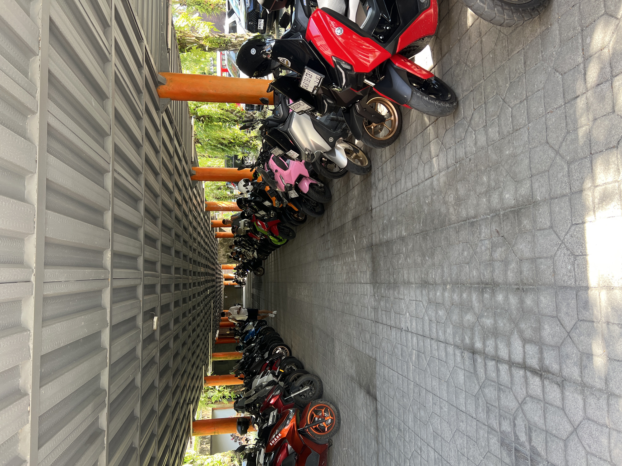 The image depicts a parking area in Petulu, Indonesia, densely packed with scooters of various colors and models. The parking area is covered by a gray corrugated metal structure supported by orange beams. A person wearing a white shirt and dark shorts is visible walking down the center of the parking area. The ground is paved with gray stone tiles. The scooters are lined up in rows, filling the parking space. Foliage is partially visible between the support beams of the parking structure.