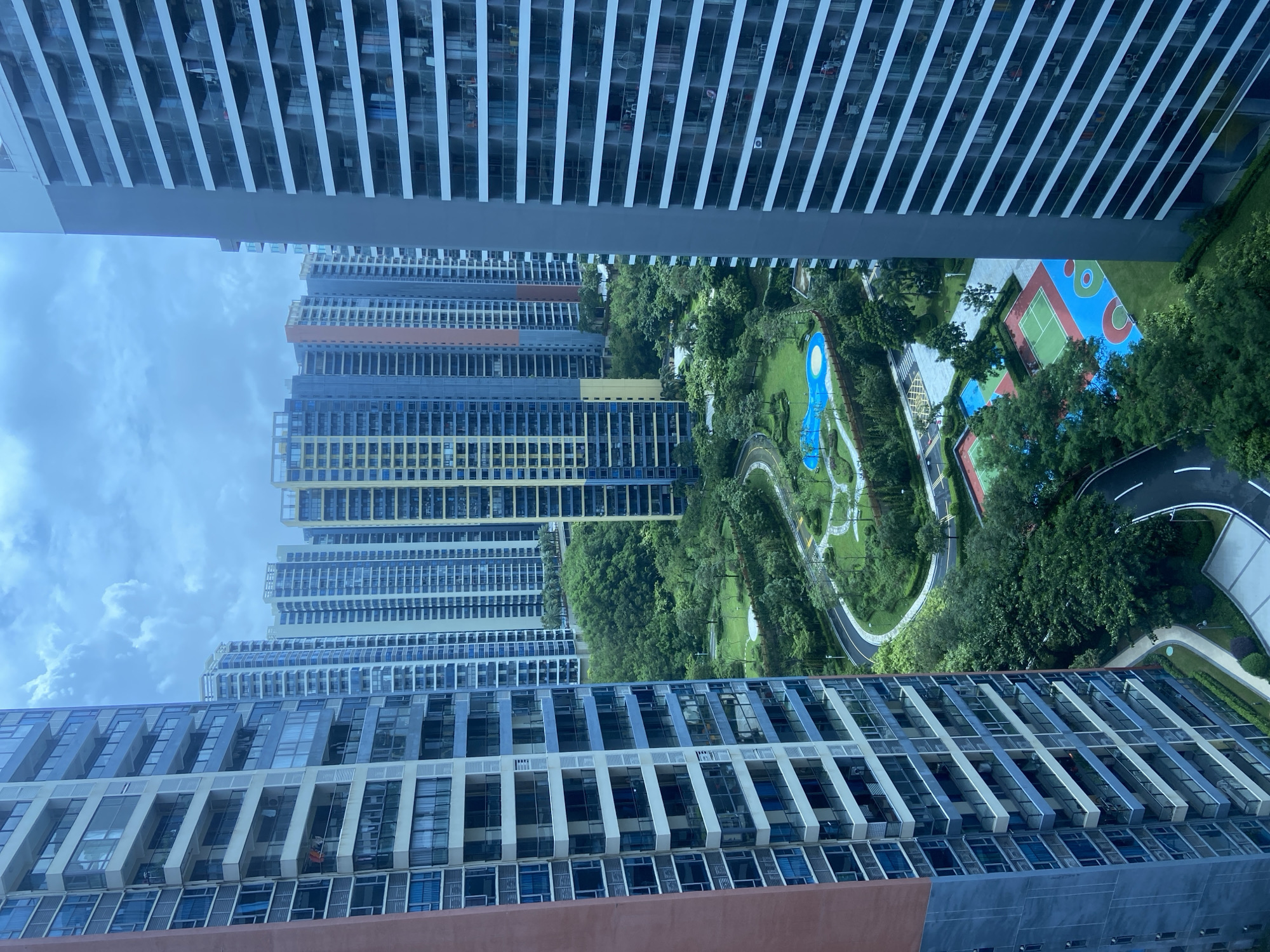 The image shows a view of a residential complex in Huizhou, China, likely in the Daya Bay district, characterized by a cluster of high-rise apartment buildings surrounding a green park area. The buildings are modern, with many floors and balconies, and vary in color, featuring shades of blue, yellow, and grey. The park below includes walking paths, grassy areas, trees, a swimming pool with a distinctive shape, and sports courts, one of which is brightly colored with red, blue, and green sections, and another is green with red accents. The sky is cloudy, suggesting an overcast day. The perspective suggests the photograph was taken from a window or balcony of one of the apartment buildings, looking out over the complex. There are no identifiable people or specific actions occurring, presenting a static urban landscape scene.