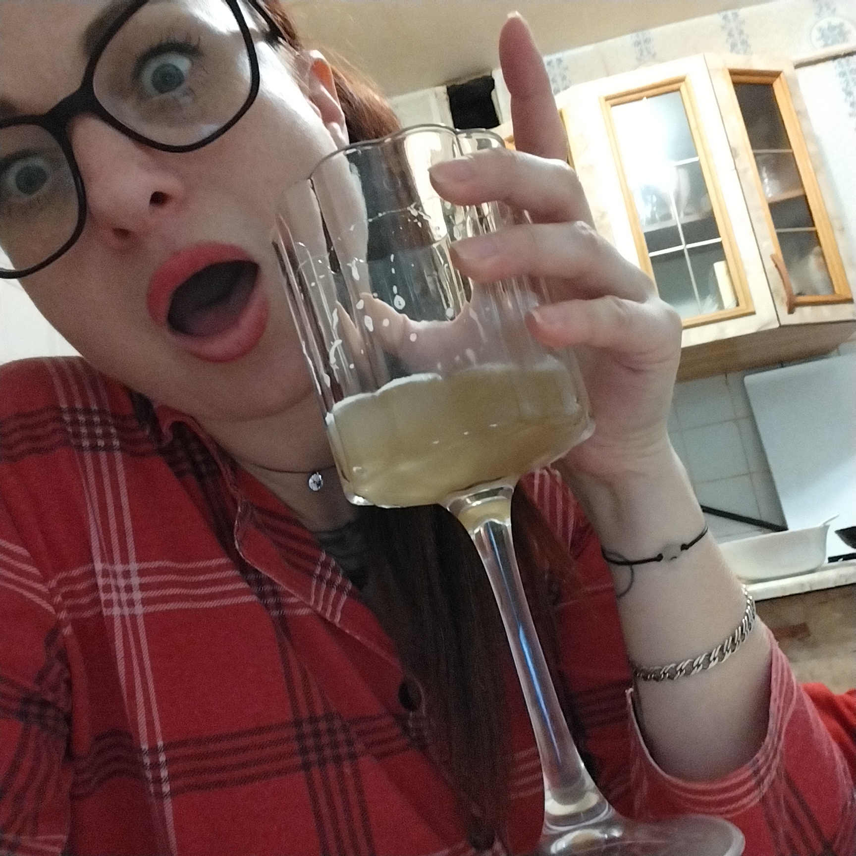 An extreme close-up shot captures a woman with wide eyes and an open mouth holding a champagne flute filled with a pale yellow liquid, possibly beer or champagne, with a frothy head. She is wearing large black-rimmed glasses, a red plaid shirt, and a delicate silver necklace. Her right hand, which is holding the glass, has a black string bracelet with a small white charm. A chunky silver bracelet adorns her left wrist. The background suggests a kitchen setting, with wooden cabinets and white tiled walls visible. The lighting is diffused, creating soft shadows.