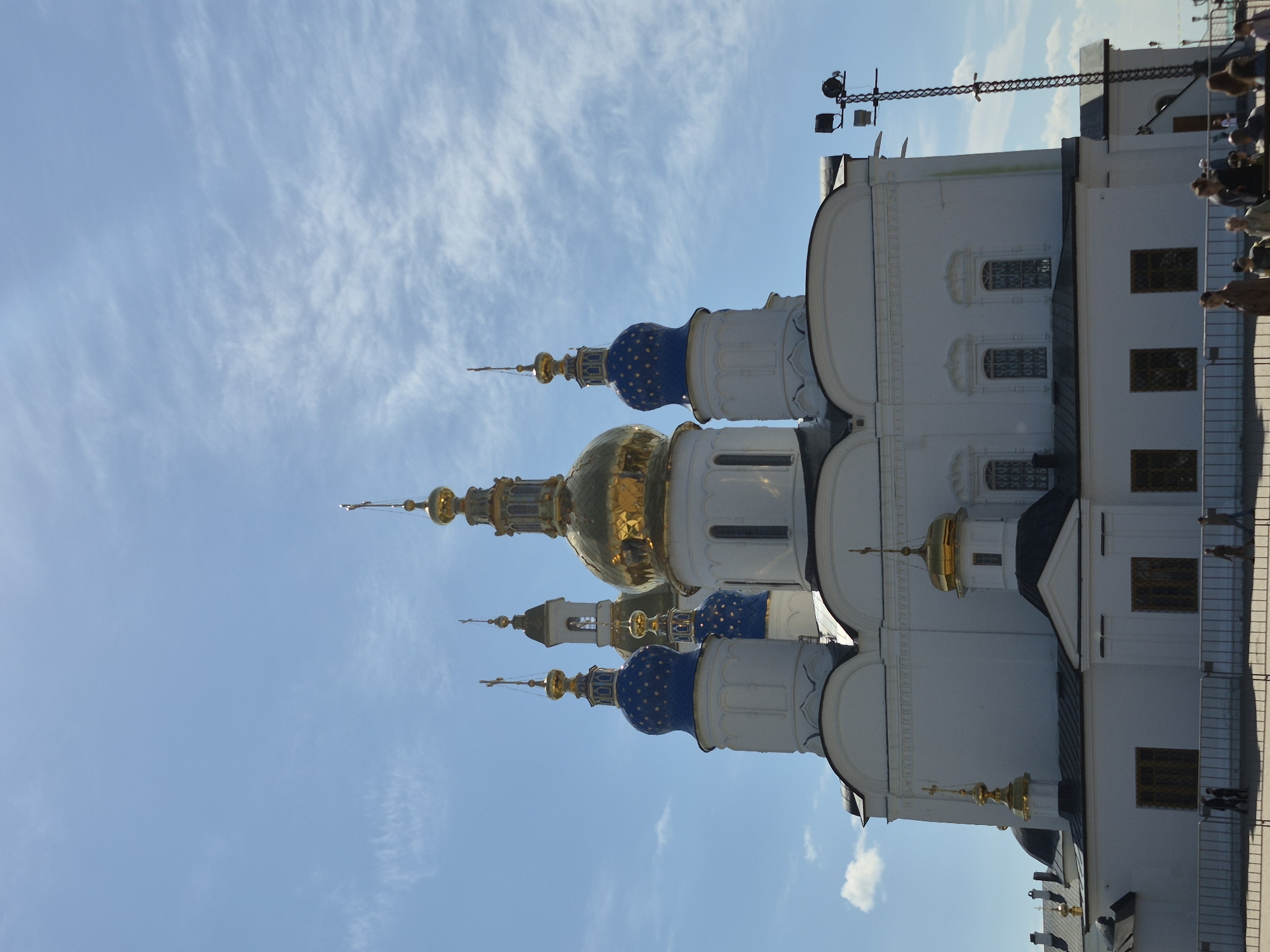 The image captures the architectural details of the Tobolsk Kremlin, specifically the domes and structures of one of its cathedrals, in Tobolsk, Russia. The image is taken at an upward angle, emphasizing the blue and gold domes with intricate detailing. The domes rise against a light blue sky with wispy white clouds. The walls of the cathedral are white, with architectural features such as arches, windows, and trim details. The presence of fencing and people suggests a public space, likely a viewpoint or pathway near the cathedral. A tall pole with lighting equipment is visible on top of the building. The overall scene captures the historical and architectural grandeur of the Tobolsk Kremlin.