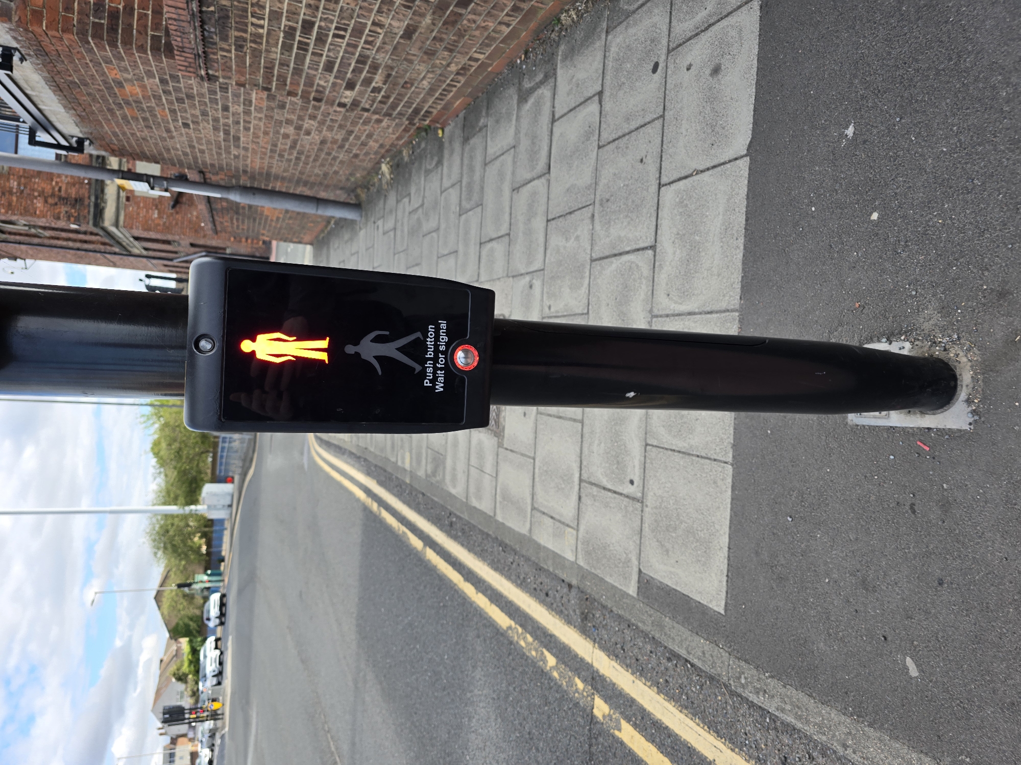 The image shows a pedestrian crossing signal post on a street corner. The signal itself displays a yellow walking person icon, indicating that it is safe for pedestrians to cross. Below the walking icon is the text "Push button, Wait for signal" and a red circular button that appears to be illuminated. The post is black and cylindrical, and it is situated on a paved sidewalk next to a street with double yellow lines. In the background, there are buildings, trees, and cars on the road, suggesting an urban environment. The sky is cloudy. The location appears to be Stockton-on-Tees, United Kingdom, based on the style of the pedestrian crossing signal and the general urban landscape.