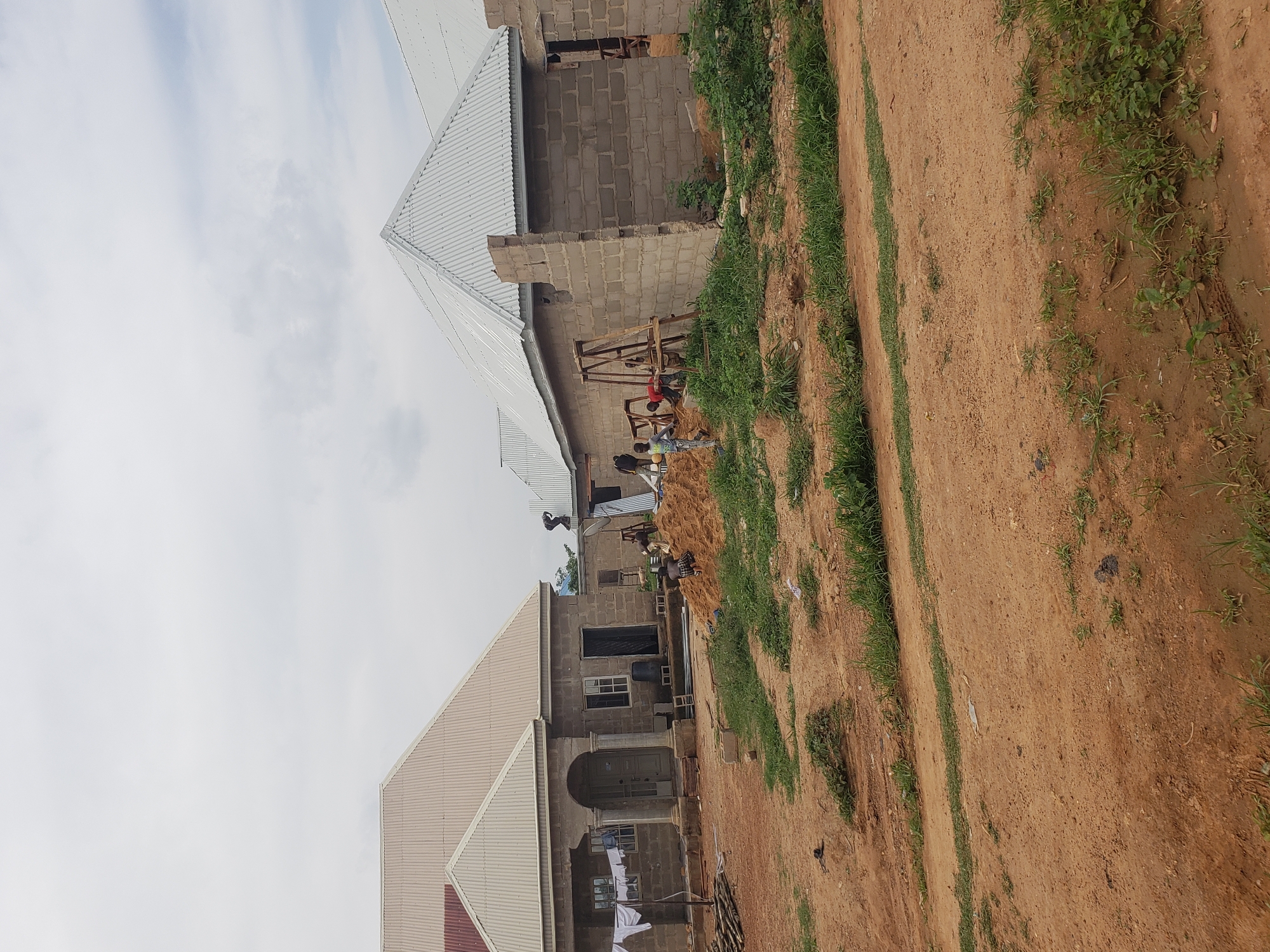 The image depicts an outdoor scene in the city of Kachia, Nigeria, featuring residential construction. In the foreground, a dirt path with scattered green grass and some debris leads towards two newly constructed houses. The house on the left is more complete, with a finished, light-colored corrugated metal roof, visible columns and archways at the entrance, and windows. Laundry is hanging on a line in front of this house. The house on the right is still under construction, with walls made of grey cinder blocks and a similar corrugated metal roof in place. Several people are engaged in construction work. One person is working on the roof of the house on the right. Others are gathered around a pile of sand near the house on the right, possibly involved in mixing or transporting materials. The environment is characterized by a cloudy sky, dry earth, and sparse vegetation.