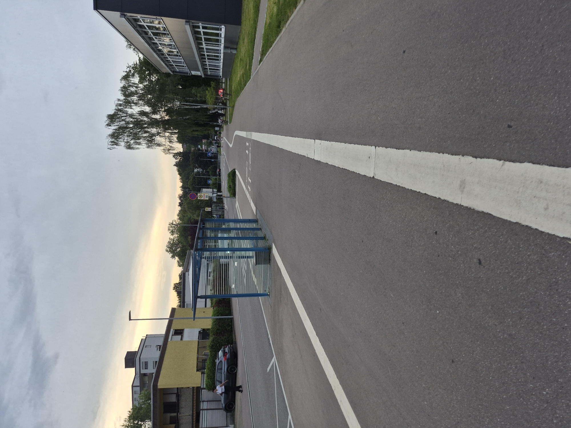The image depicts a street scene in an urban environment, likely in the late afternoon or early evening, indicated by the overcast sky and soft lighting. In the foreground, a paved road with a white line down the center is visible, suggesting a clear division of lanes. To the left of the road, a modern bus stop with a glass enclosure and blue metal framing stands. Adjacent to the bus stop, a dark-colored car is parked, and a person in dark clothing is walking away from it towards the bus stop.
Further in the background, a multi-story building with a yellow facade and balconies is situated. To the right, a dark-colored building with numerous large windows runs parallel to the road. Beyond these buildings, lush green trees and vegetation are abundant. The overall scene suggests a well-maintained residential or commercial area with dedicated infrastructure for public transportation and cycling. The specific location is identified as Unknown, Unknown.