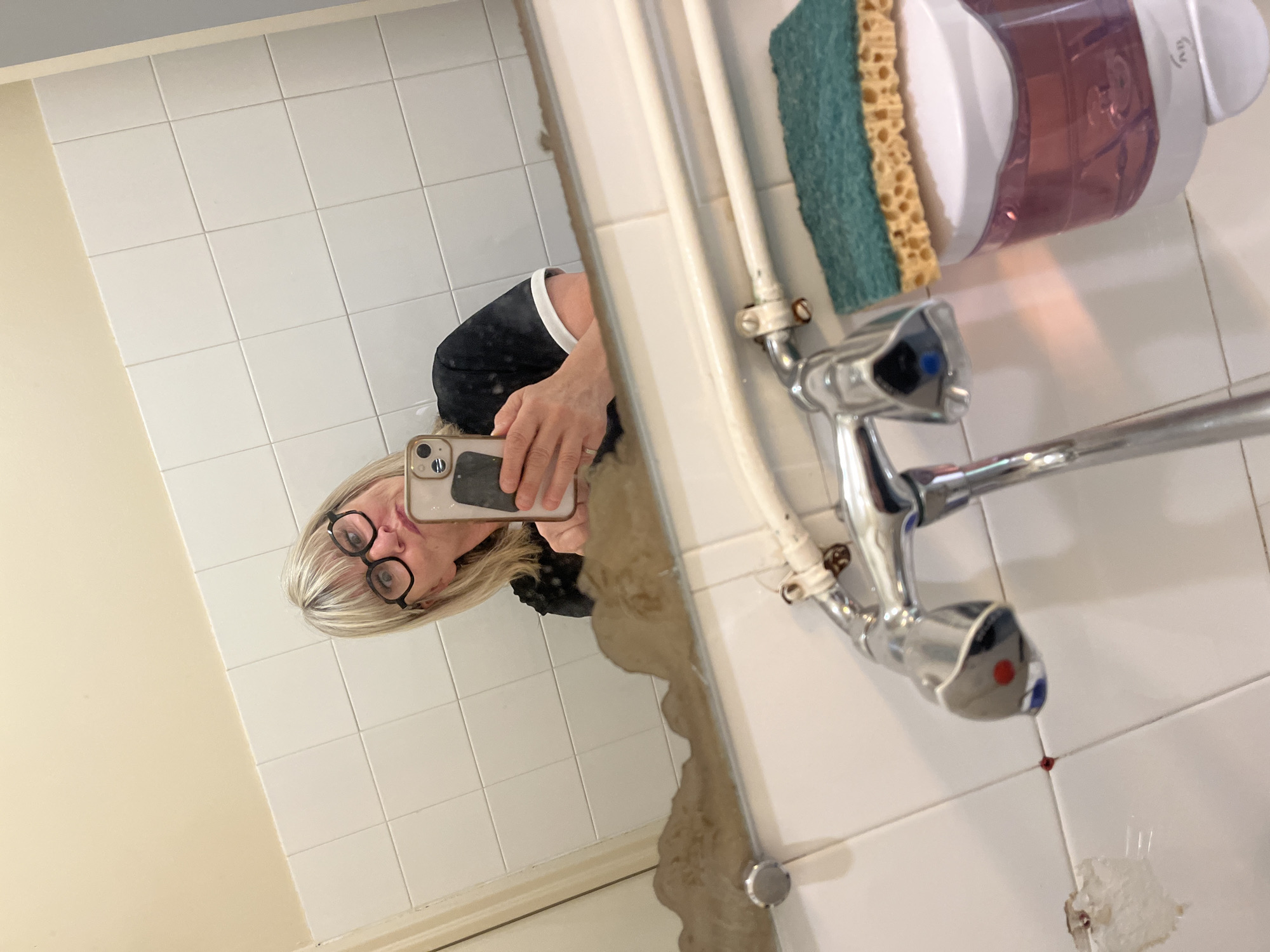 In La Fouillouse, France, a person with blonde hair and glasses is taking a selfie in a bathroom mirror. The person is wearing a black shirt with a white collar. The mirror reflects a sink with a chrome faucet, a soap dispenser, and a sponge. The bathroom walls are tiled with white squares. The sink area shows some wear and tear, with peeling grout and water stains. The lighting is artificial, creating reflections on the shiny surfaces.