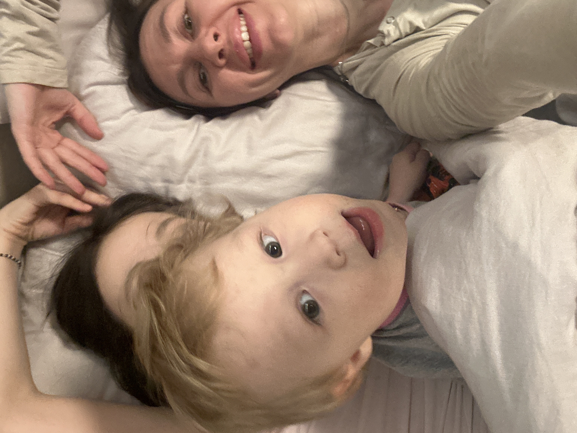 This media file captures a close-up, top-down view of an adult woman and a young child lying together, likely on a bed or soft surface. Both subjects are looking upwards towards the camera, suggesting it may be a selfie.
The adult woman, positioned in the upper portion of the frame, has dark hair and is smiling broadly, revealing her teeth. She is wearing a light-colored, possibly beige or light grey, long-sleeved top. Her right hand is visible, resting gently near the child's head.
The young child, located in the lower part of the frame, has light blond or light brown hair and wide, curious dark eyes. Their mouth is slightly open. The child is wearing a grey top, with a hint of pink visible around the collar area.
Both individuals appear happy and content, with the woman displaying a clear smile and the child looking engaged. They are situated on a light-colored, textured fabric, likely bedding, which dominates the background and indicates an indoor setting. The lighting is soft and even. There is no visible text within the image. The location context for this media is Votkinsk, Russia.
