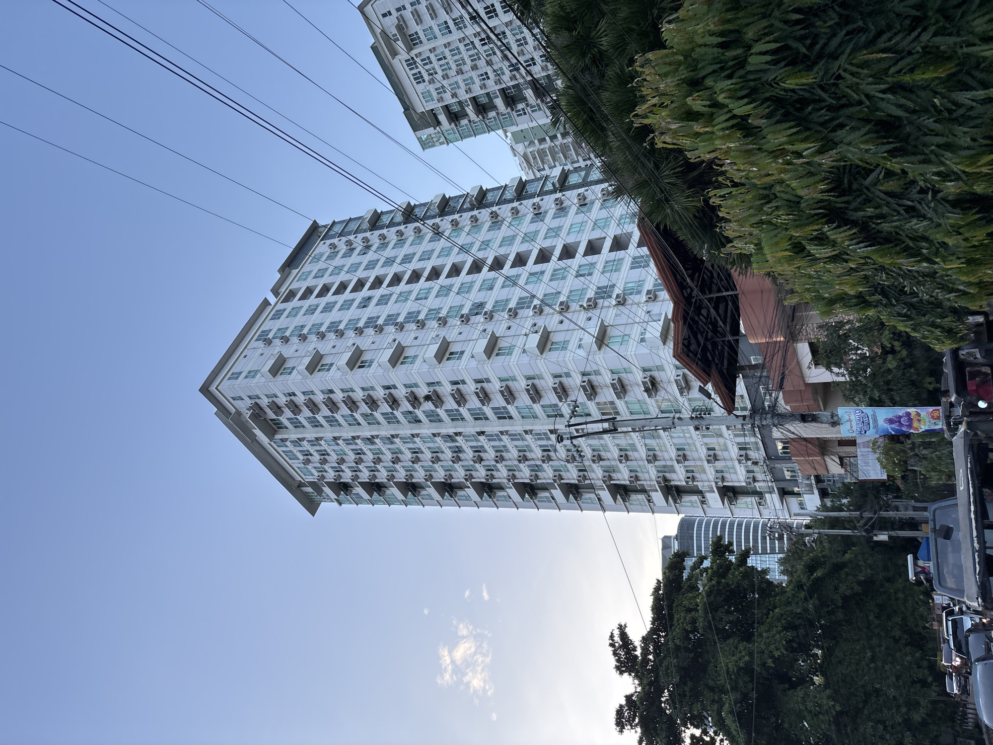 The image shows a view of a street in Davao City, Philippines, with several apartment buildings and trees. The primary focus is a tall, white apartment building with many balconies and air conditioning units. In the background, another apartment building is visible, as well as a modern-looking curved glass building. Power lines are prominent in the foreground, crisscrossing the sky. Dense green foliage from trees partially obscures the lower part of the scene. Several vehicles, including SUVs and cars, are on the street. A billboard for "Halo-halo Special" is also visible, advertising a popular Filipino dessert. The sky is a clear blue with a few wispy clouds.