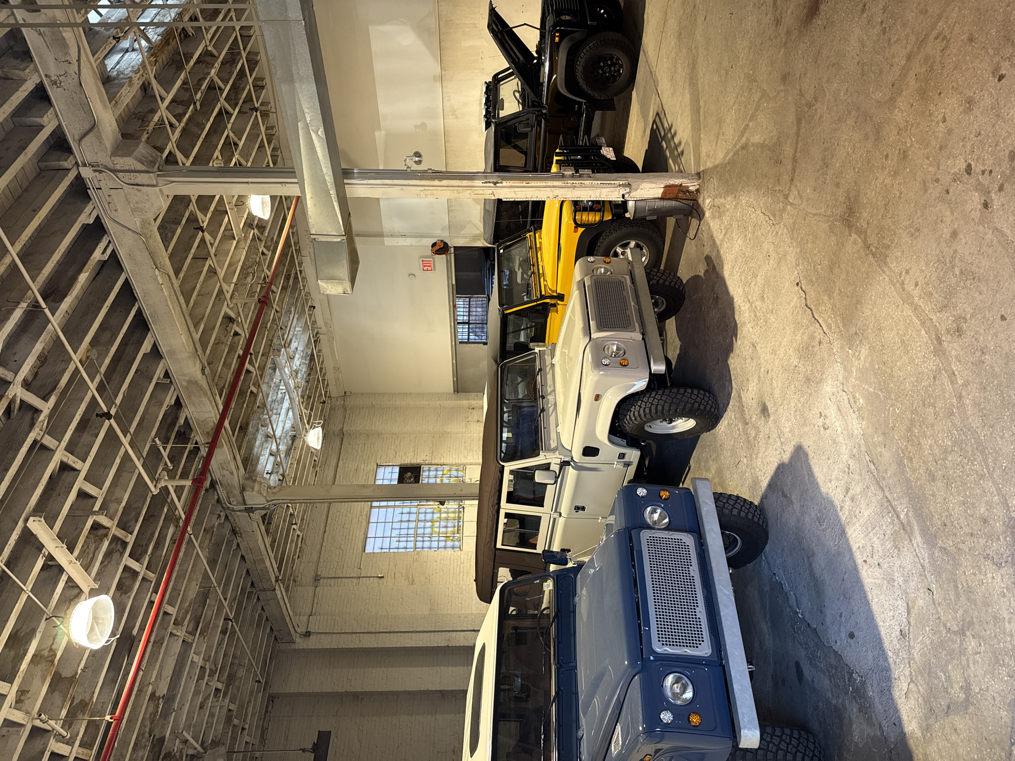 The image depicts the interior of a large industrial space, likely a warehouse or a garage, in New York City, United States of America.
The space is characterized by high ceilings with exposed beam construction, including a network of steel and wooden supports. Several large windows are visible along one wall, indicating the presence of natural light. The walls appear to be made of painted brick. The floor is concrete.
A line of four Land Rover Defender vehicles are parked in the foreground, oriented along the length of the space. The vehicles range in color from dark blue to white to yellow to black.
Artificial lighting is provided by suspended lamps. A red fire suppression pipe runs along the ceiling structure. There is an exit sign visible near the center of the image. No people are visible.
