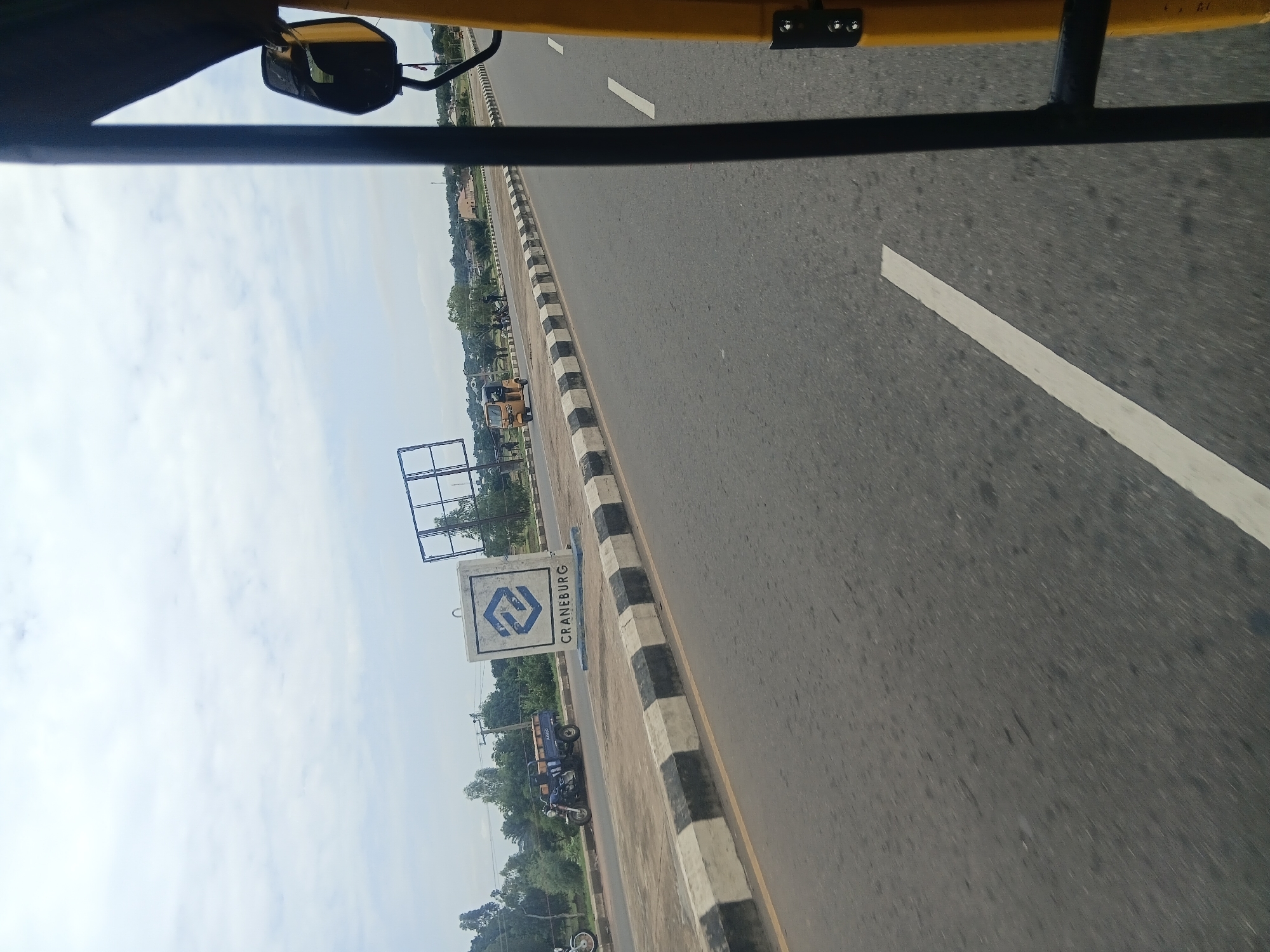 The image captures a road scene in Jalingo, Nigeria, taken from the perspective of a vehicle. A yellow and black three-wheeled vehicle is partially visible in the foreground on the right side of the frame, indicating the viewpoint. The road itself is paved with asphalt, marked with white dashed lines. A black and white striped concrete barrier runs along the left side of the road.
To the left of the road, a sign with a blue logo and the text "CRANEBURG" is visible on a white rectangular base. Next to the sign, a blue three-wheeled vehicle with a cargo bed is parked. Further down the road, another yellow three-wheeled vehicle is in motion. In the background, there are trees, buildings, and a large, empty billboard structure. The sky is overcast with clouds. The scene suggests a commercial or roadside area within the city.