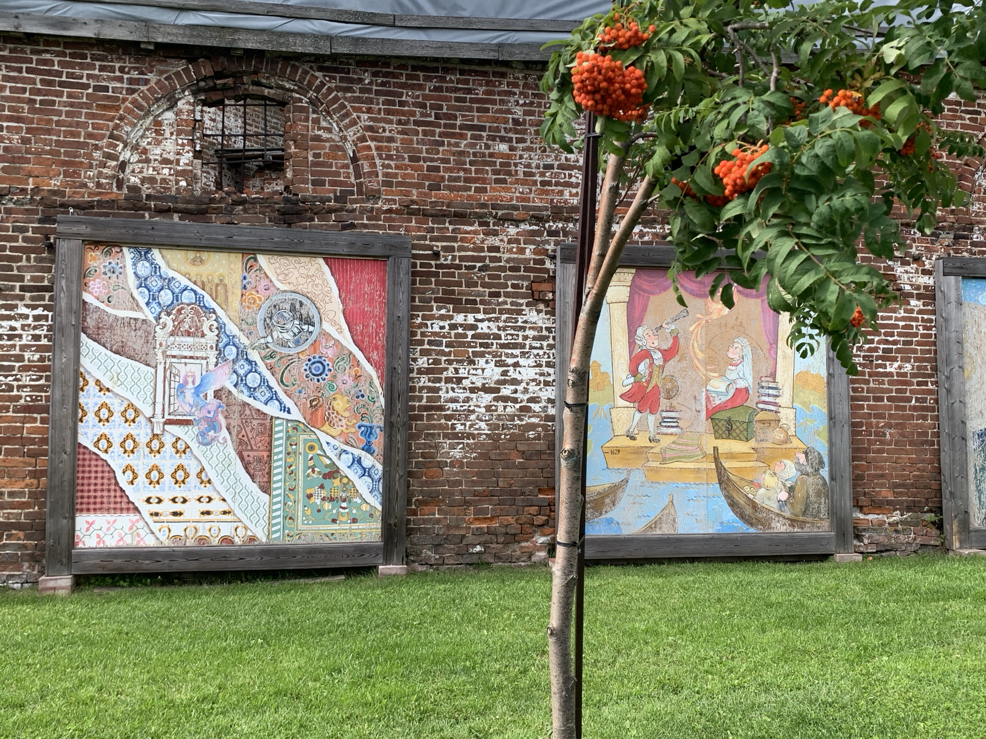 The image shows a section of a weathered red brick wall in Usolye, Russia, with two large, framed artworks displayed in front of it. To the left, the artwork is a colorful mosaic or collage composed of various patterns and images, including what appears to be tiles and painted sections. A circular element with a metallic sheen is prominent in the upper right portion of this piece. To the right, a second framed artwork depicts a scene with figures in historical clothing. One figure, a man in a red coat, is holding a spyglass and standing on a boat. Another boat contains people, and books are visible on a surface in the background. A date, "1629", is inscribed on this artwork. A young rowan tree with bright orange berries stands in the foreground, partially obscuring the artworks and the brick wall. The ground in front of the artworks is covered in well-maintained green grass. The brick wall shows signs of age, with some mortar missing and bricks discolored. An arched opening in the wall, partially covered by a metal grate, is visible above the left artwork.
