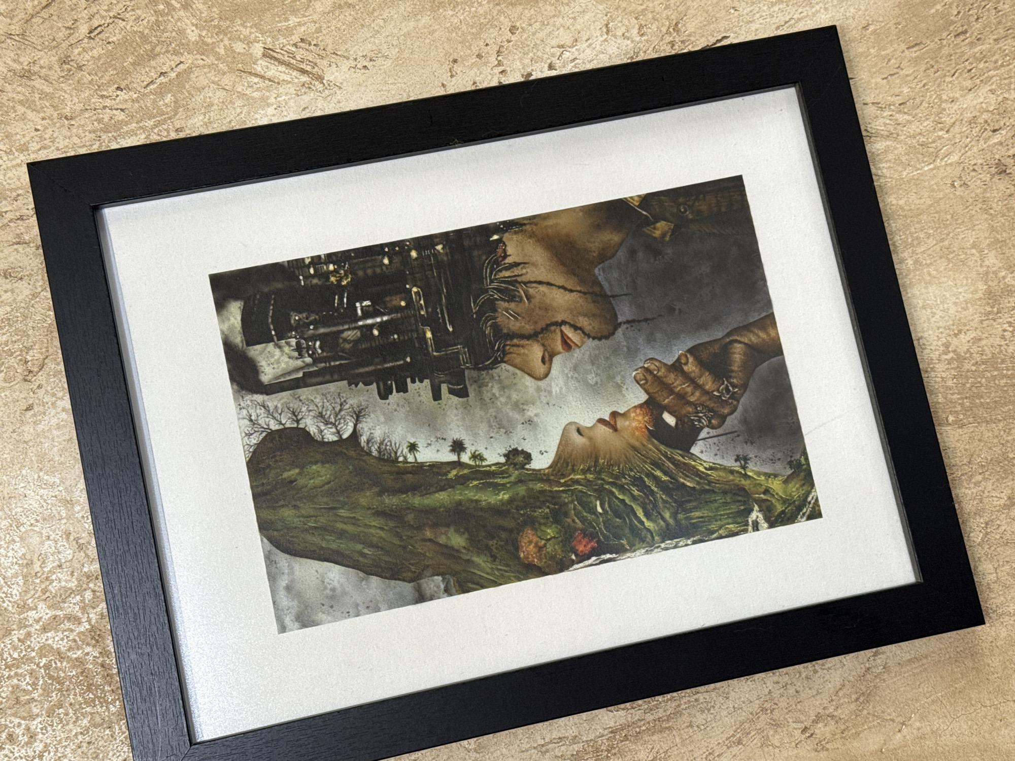 A framed print rests on a surface. The print depicts a face constructed with machinery above and a face constructed of earth below, both facing each other. The frame is black and surrounds a white mat. The surface is beige and textured.