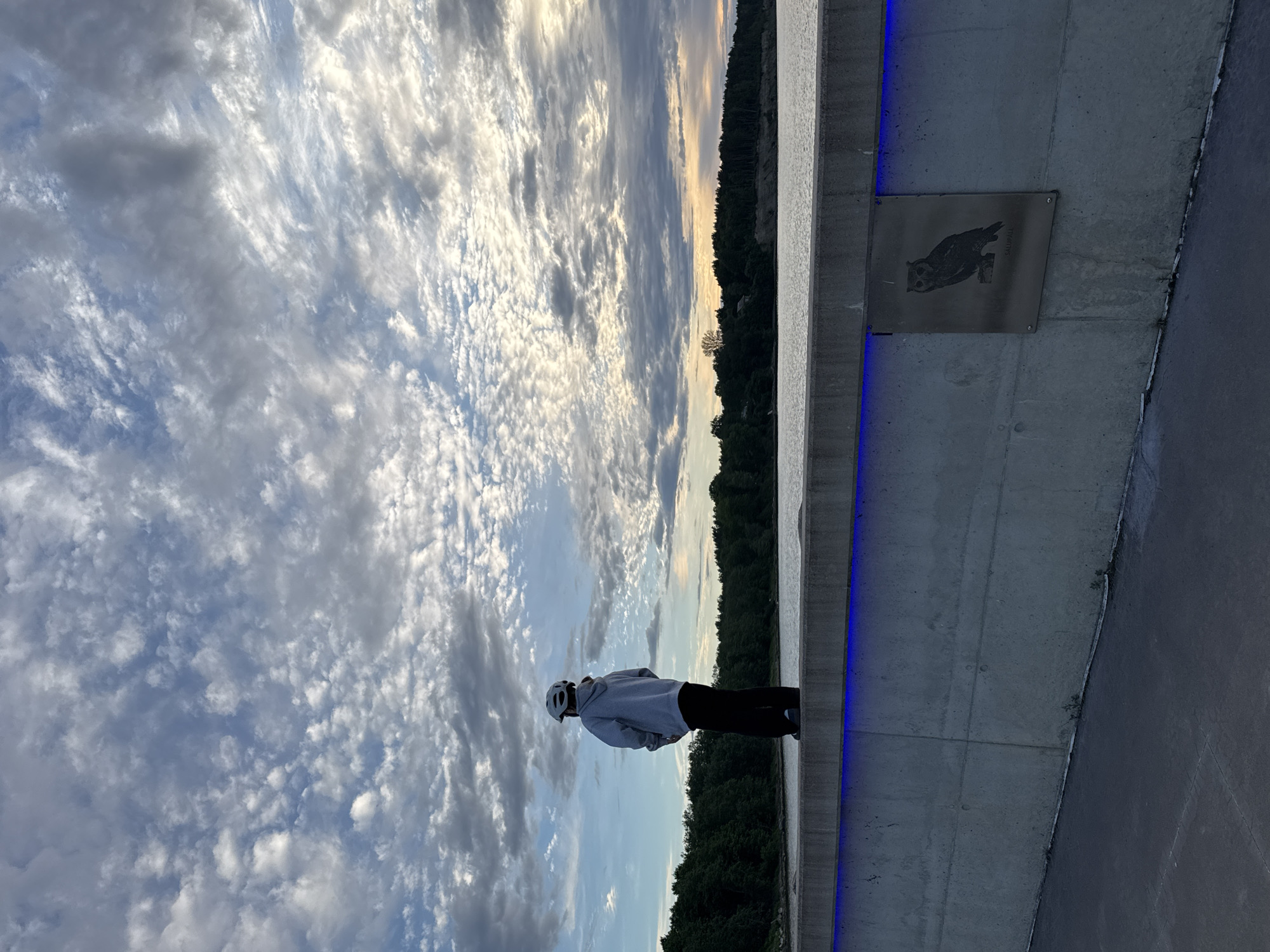 A person wearing a grey hoodie, black pants, and a white helmet stands on a concrete wall, facing away from the camera and looking out at a body of water. The wall is illuminated with blue lights along its top edge. On the wall, to the right of the person, is a metal plaque with an etched image of an owl and text that reads "SALUPALL". The water extends to a tree-lined shore in the distance, under a sky filled with dramatic, scattered clouds. The scene appears to be a waterfront promenade or embankment. The location is the Pirita promenade in Tallinn, Estonia.