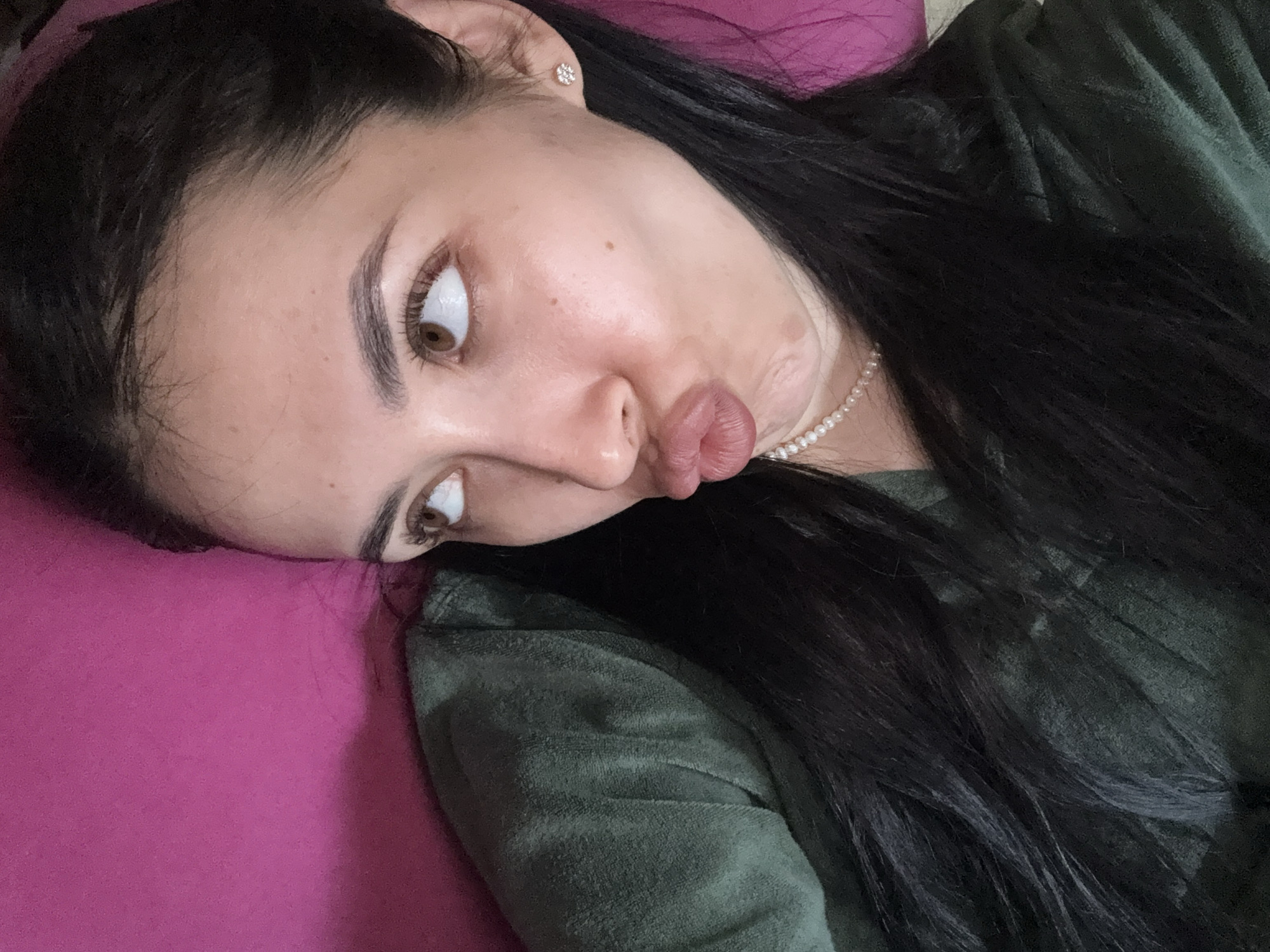 A woman with dark hair and brown eyes is lying on a pink pillow. She is wearing a green top and a pearl necklace. She has a playful expression with pursed lips. The image does not provide any information about the location being Votkinsk, Russia, or any other details about the environment.