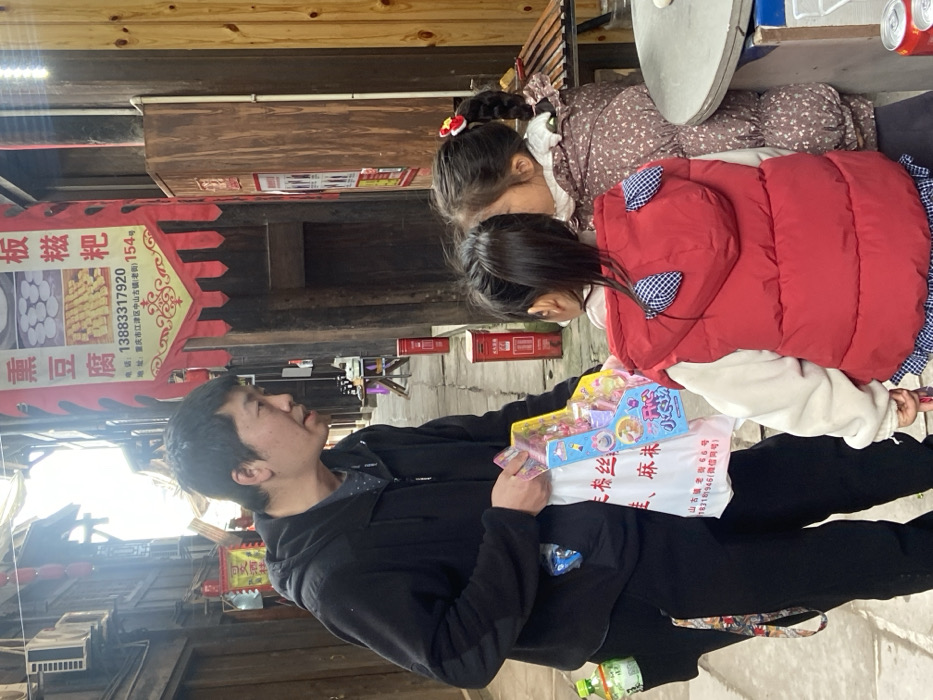 An outdoor scene depicts a man in a black jacket holding toys in a white bag. Two young girls are nearby, one in a floral dress and the other in a red vest. A banner advertises local food items, including "Ban Ci Ba" (rice cake) and smoked tofu. The streetscape features traditional Chinese architecture, including wooden buildings and red mailboxes. Text on the banner shows a phone number and address in Chongqing.