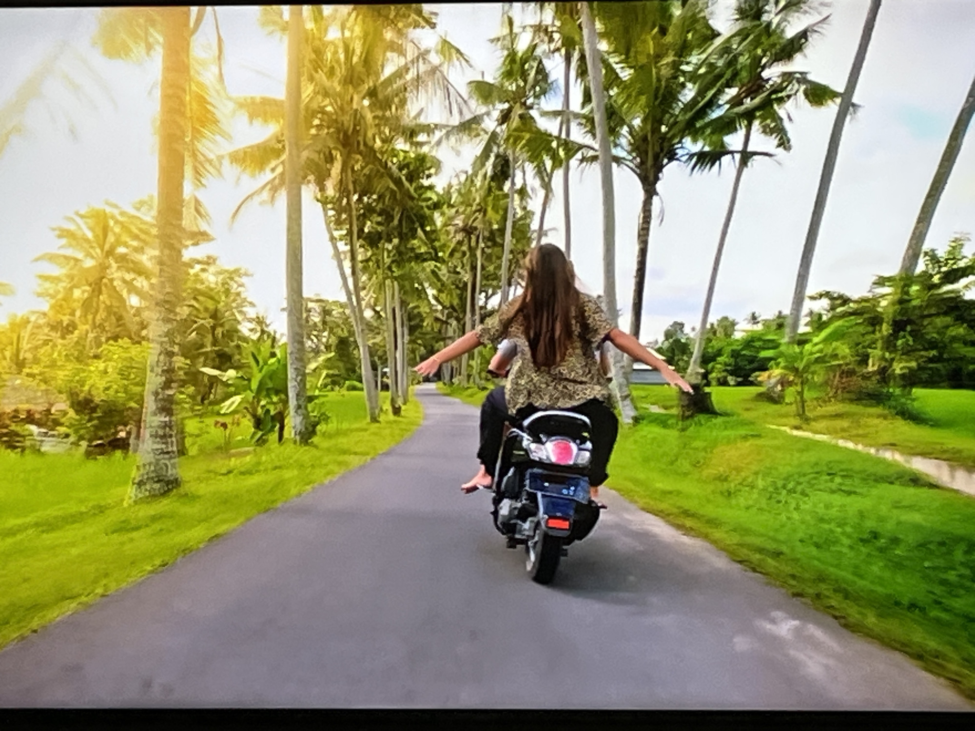 Two individuals are riding a scooter down a paved road lined with palm trees. The person in front is driving the scooter, while the person behind is seated facing forward. Both individuals are wearing casual clothing. The surrounding environment appears to be tropical, with lush green vegetation and clear skies. The lighting suggests it is daytime, possibly late afternoon due to the warm, golden hues of the sunlight filtering through the palm trees. The road is asphalt and curves slightly to the left. The scene is peaceful and scenic, evoking a sense of travel or vacation. There is no indication of this scene being located in Kharkiv, Ukraine.