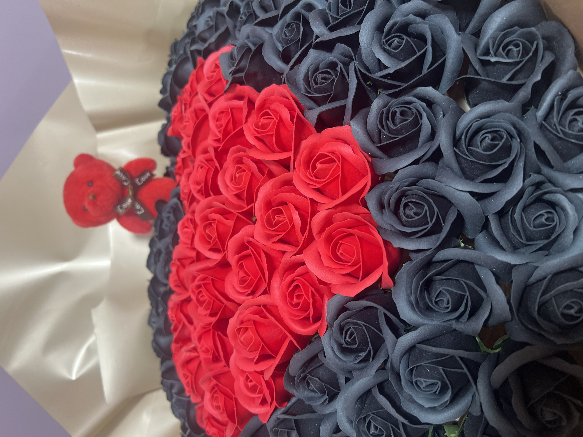 The image shows a bouquet of roses, predominantly black, with a heart shape formed by red roses in the center. A small red teddy bear with a ribbon that says "Love" is attached to the wrapping of the bouquet. The bouquet is wrapped in a light-colored paper. The background appears to be a plain wall. There is no evidence indicating the location is Kavarna, Bulgaria.