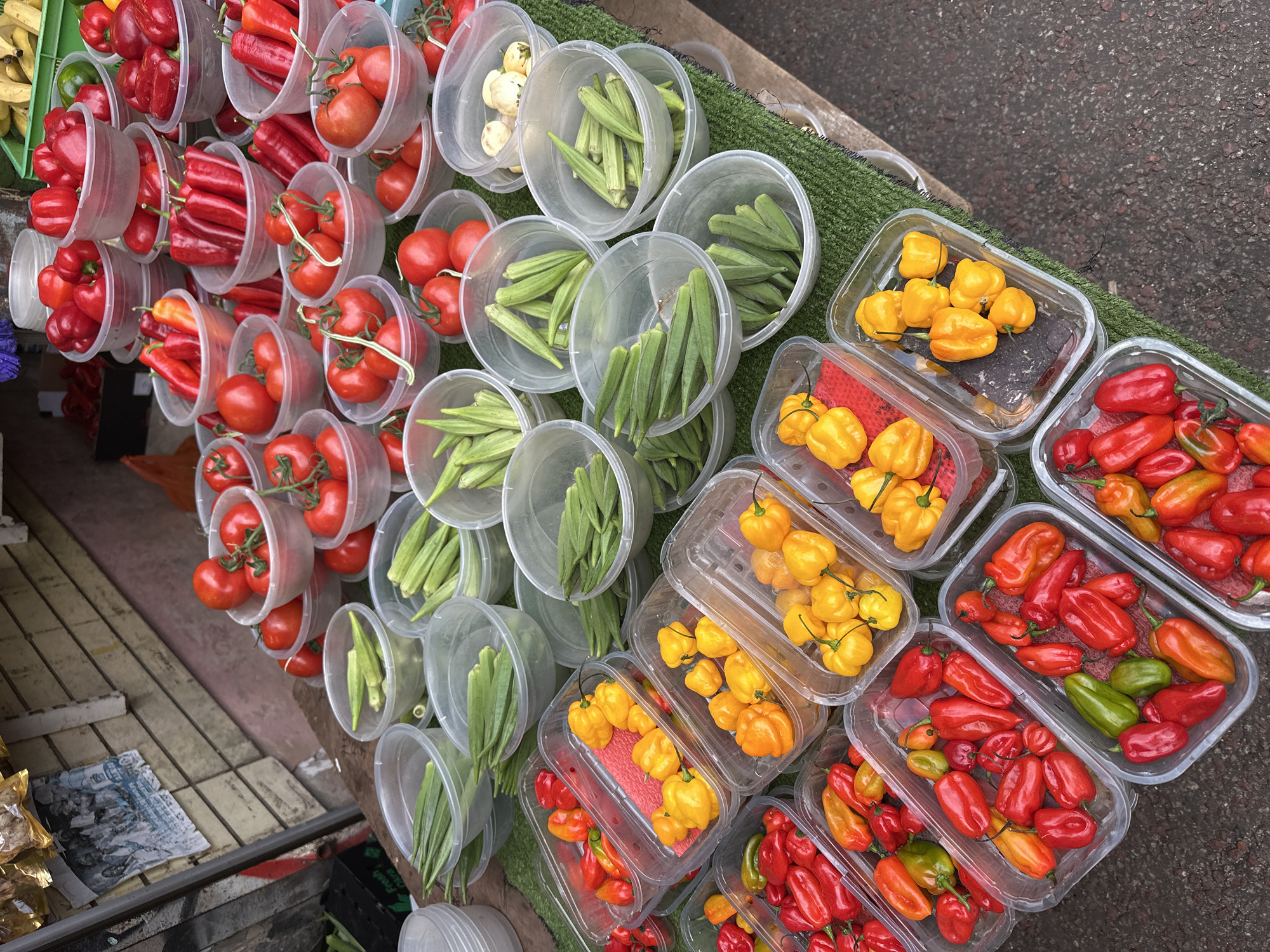 A high-angle shot shows a street-market display of produce in plastic containers. On the left are round containers holding red bell peppers, red tomatoes on the vine, and green okra. To the right, rectangular containers hold yellow and red scotch bonnet peppers. The display is set on a green astroturf mat, resting on a table. To the far right, the edge of a paved street is visible. In the lower left corner, there is a partial view of what appears to be a lower shelf or surface with a newspaper. This scene is probably a street market somewhere in London, United Kingdom.