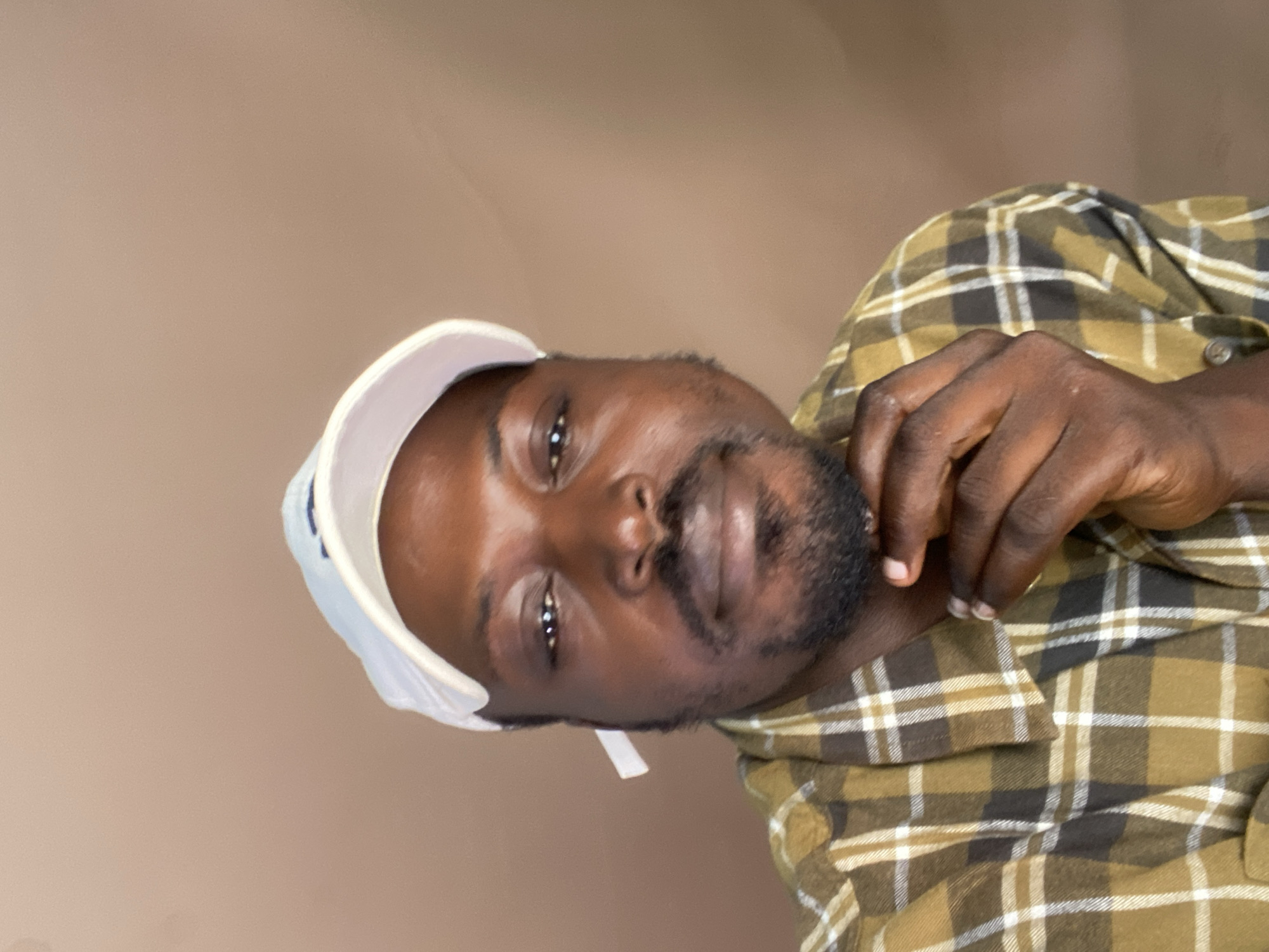 A medium shot shows a Black man wearing a light-colored baseball cap and a plaid shirt. The man is looking directly at the camera with his chin resting on his hand. He has a beard and mustache. The background is a plain, light brown color. There is no indication of the location being in Gura Top, Nigeria.
