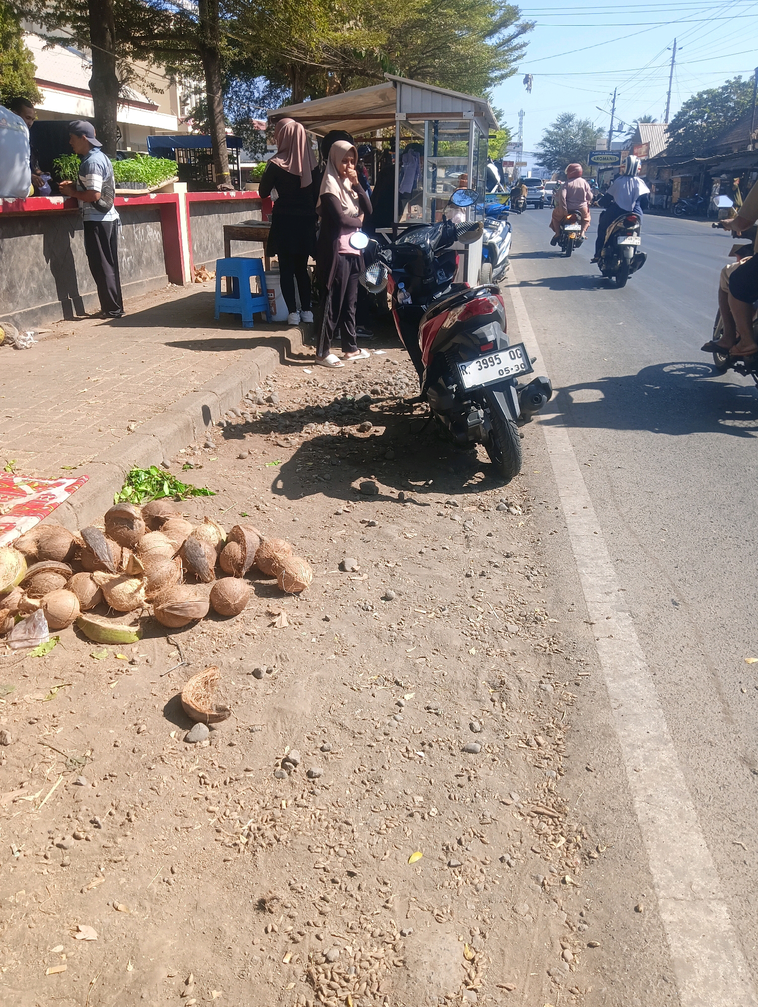 The image depicts a street scene in Sokaraja, Indonesia, on a sunny day. In the foreground, a pile of coconuts lies on the dusty ground near a curb. To the left, a red-and-white stall or structure is visible, with some people standing near it. One person, wearing a blue cap and striped shirt, appears to be interacting with someone at the stall.
Further down the street, several motorcycles are parked and in motion. One red motorcycle is prominently displayed in the mid-ground, with its license plate reading "R 3995 QG 05-30". A woman wearing a hijab is seated on this motorcycle. Other motorcycles and riders are seen further down the road, with some moving away from the camera. Buildings and trees line the street, suggesting an urban or semi-urban environment. The overall impression is a busy street scene with vendors and traffic.