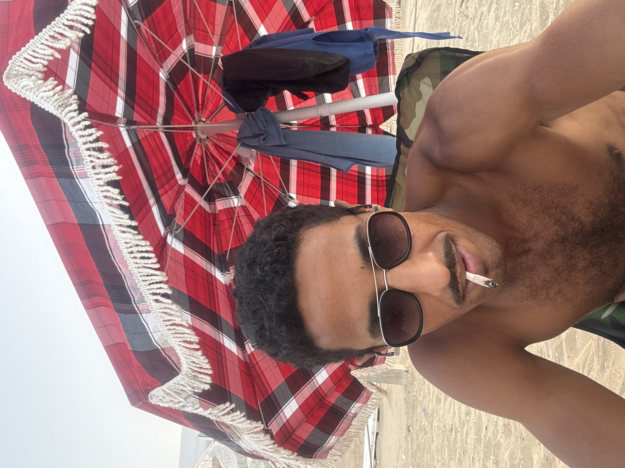 A shirtless man with a mustache and sunglasses is sitting on a beach chair under a red and black plaid umbrella. He is holding a lit cigarette in his mouth. The umbrella has white fringe around its edge. In the background, there is sand and a light-colored structure. The man is wearing a camouflage-patterned fabric over his shoulders, which is part of the chair.