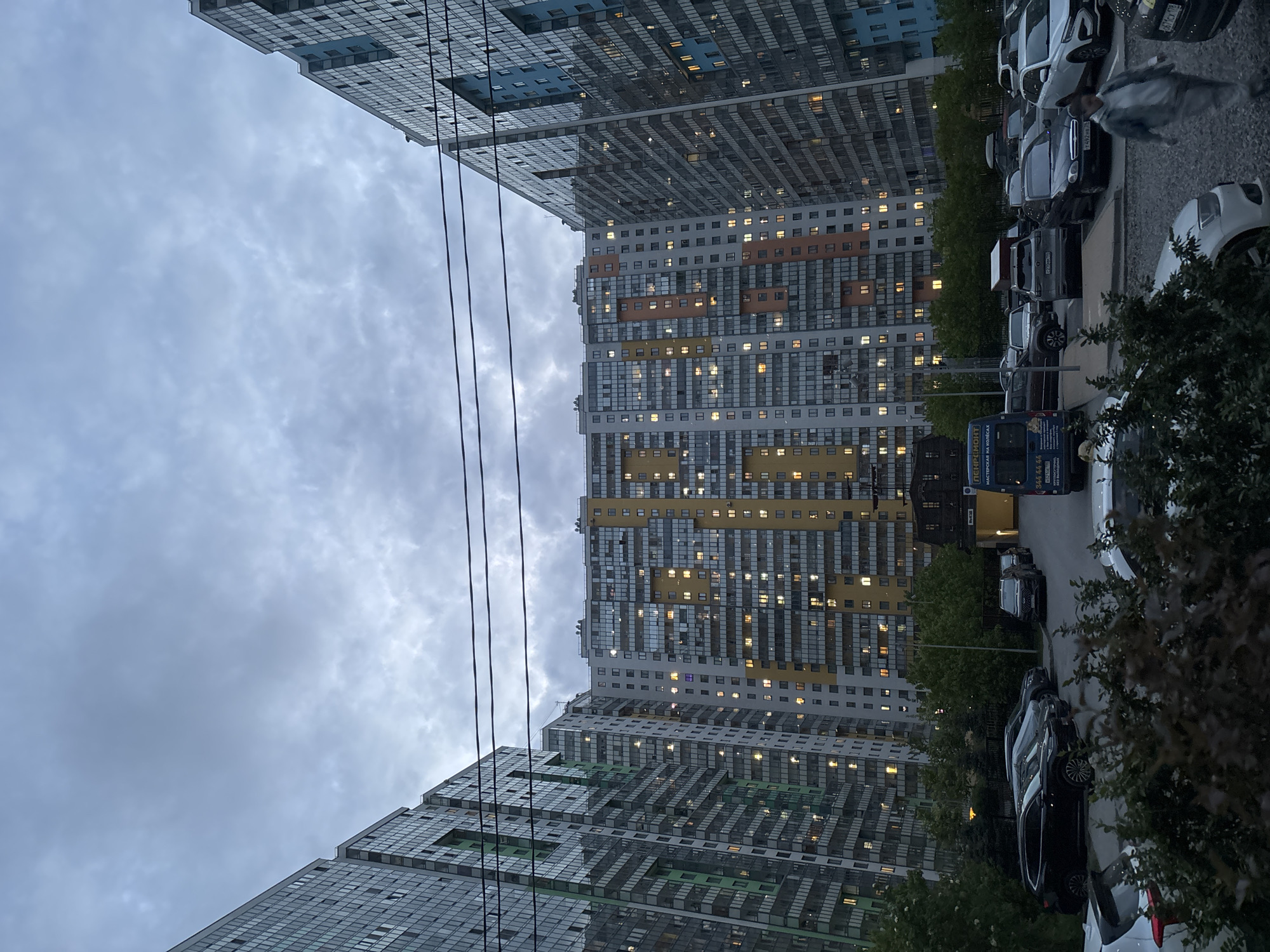 The image depicts a high-rise residential area in Saint Petersburg, Russia, under an overcast sky. Two tall apartment buildings frame a street lined with parked cars. The buildings have a modern design with many windows, some of which are illuminated, suggesting it is evening. Power lines are visible against the sky. A blue van is parked along the street. Trees are interspersed along the street and in front of the buildings.