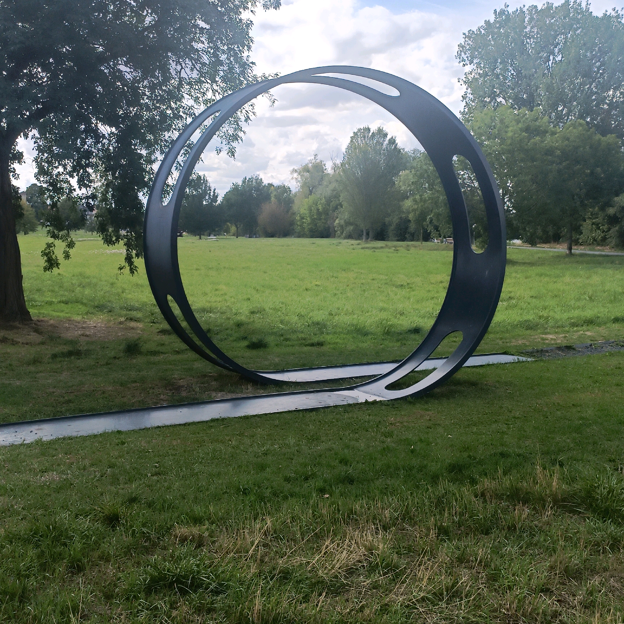 The image shows a large, dark metal sculpture in the shape of an oval ring with two internal oval cutouts, situated in a grassy park. The sculpture is partially submerged in a dark, elongated metal track that runs along the ground. The park features a large tree on the left, with sparse branches. In the background, there is a wide expanse of green grass, with a line of trees and bushes in the distance. The sky is partly cloudy. The sculpture is the central focus of the image, positioned in the foreground and mid-ground. The overall scene depicts an outdoor public space with artistic installation. This installation is located in theOpelvillen park in Rüsselsheim am Main, Germany.