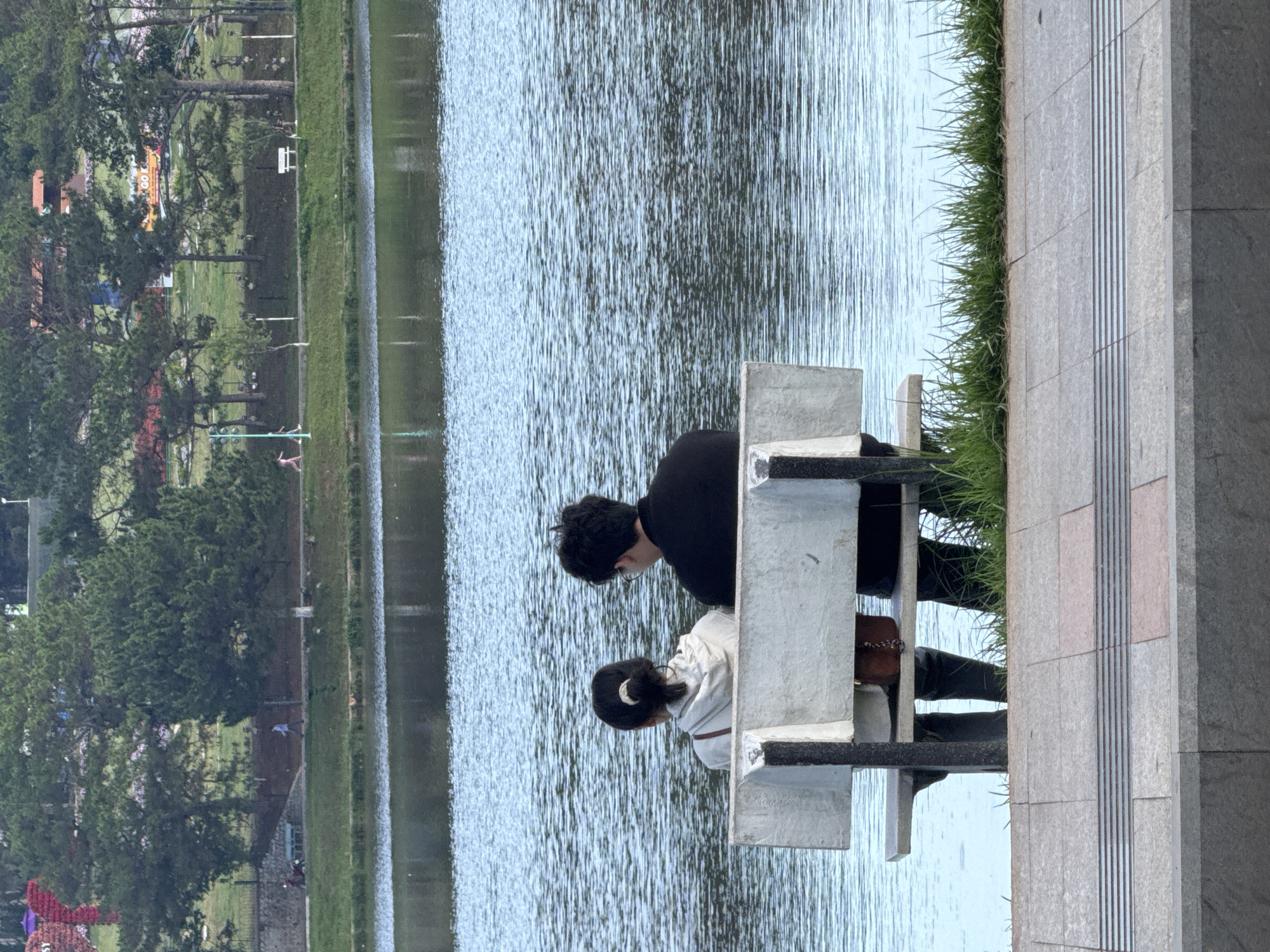 A couple sits on a stone bench beside the Xuan Huong Lake in Da Lat, Vietnam. The lake fills the center of the frame, stretching between the bench and a grassy shoreline lined with trees and buildings in the background. The couple is positioned to the right, with a man in a black shirt and a woman with her hair in a ponytail, wearing a white jacket. They both lean forward looking at something in the water. The stone bench is made of light gray blocks. Beyond the bench is a narrow strip of grass and a paved walkway with tactile paving.