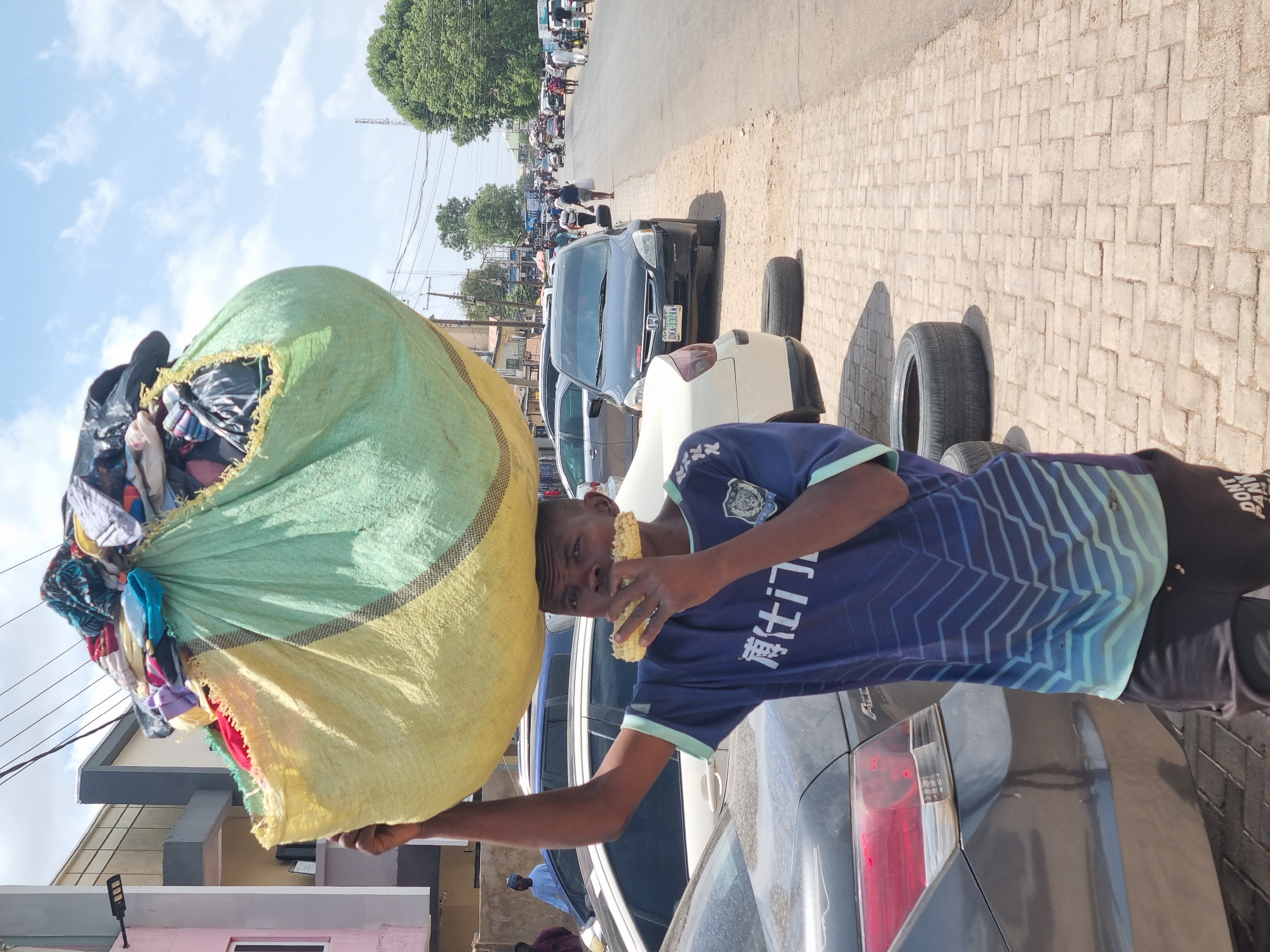 A medium shot depicts a man on a street in Jos, Nigeria. He balances a large, striped, overstuffed bag of clothing on his head and shoulder. The man is wearing a blue soccer jersey with white lettering on the sleeves and eating an ear of corn. He is positioned on a paved street lined with parked cars, including a black Honda with license plate "OKA 132AN" and a white car with a tire resting near its rear. A crowd of people can be seen further down the street. The sky is partly cloudy, with buildings and power lines visible in the background.