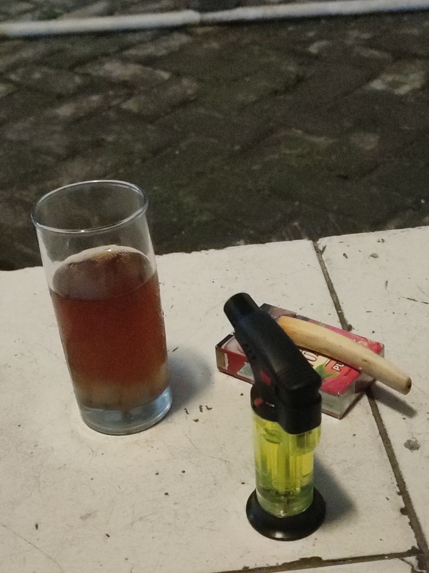 An outdoor setting at night, possibly in Jember, Indonesia, features a glass of amber-colored liquid with foam on top, a cigarette lighter, and a cigarette pack with a clove cigarette resting on top. The surface appears to be tiled pavement.