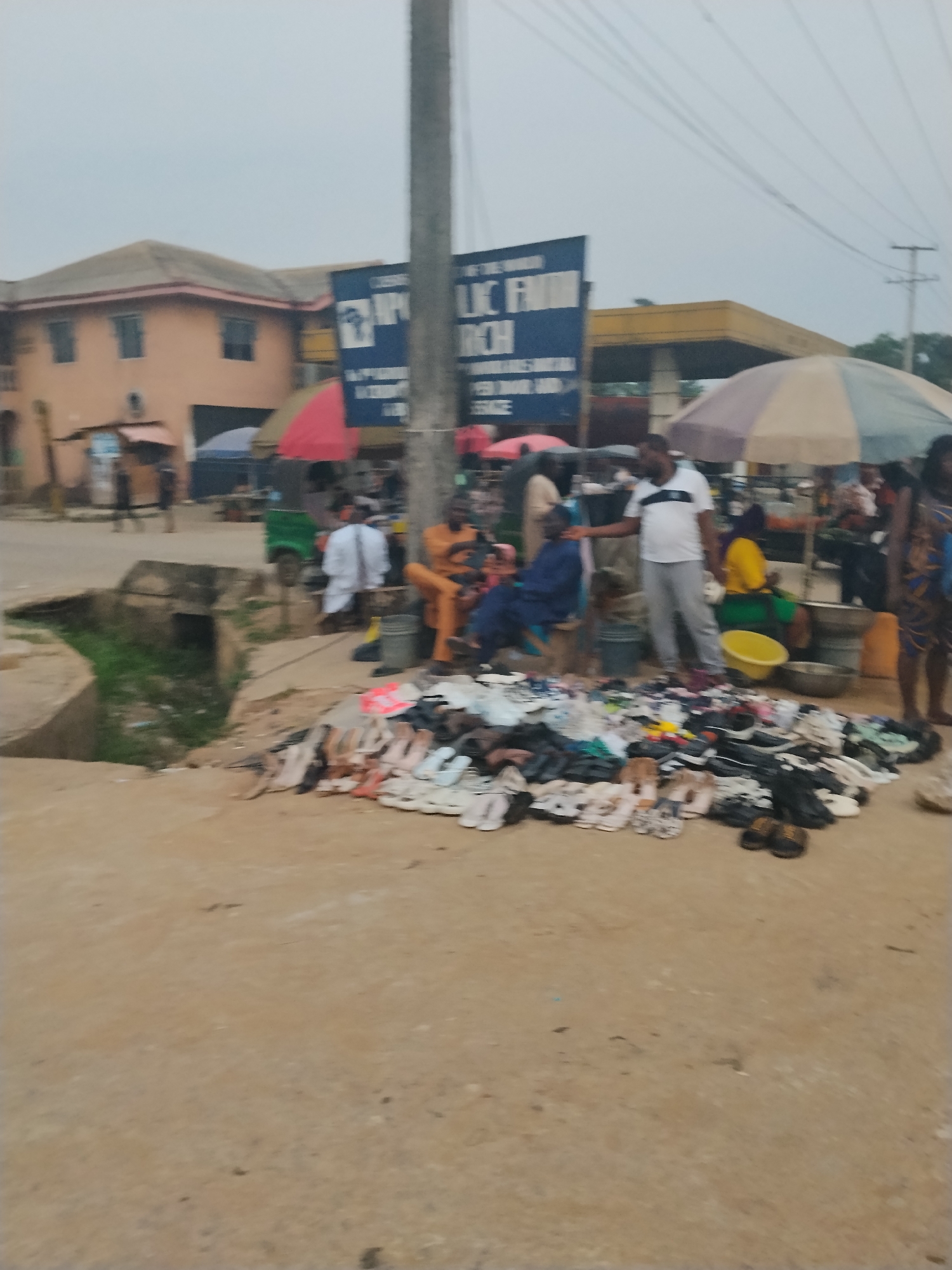 Location: Ura Nta Umuarandu, Nigeria.
Scene: A street market is active. A large collection of sandals and shoes is displayed on the ground. Several individuals are interacting with the shoe vendor. Nearby are market vendors under umbrellas. A building, a gas station, and utility poles are visible in the background. A concrete drainage ditch is in the foreground.