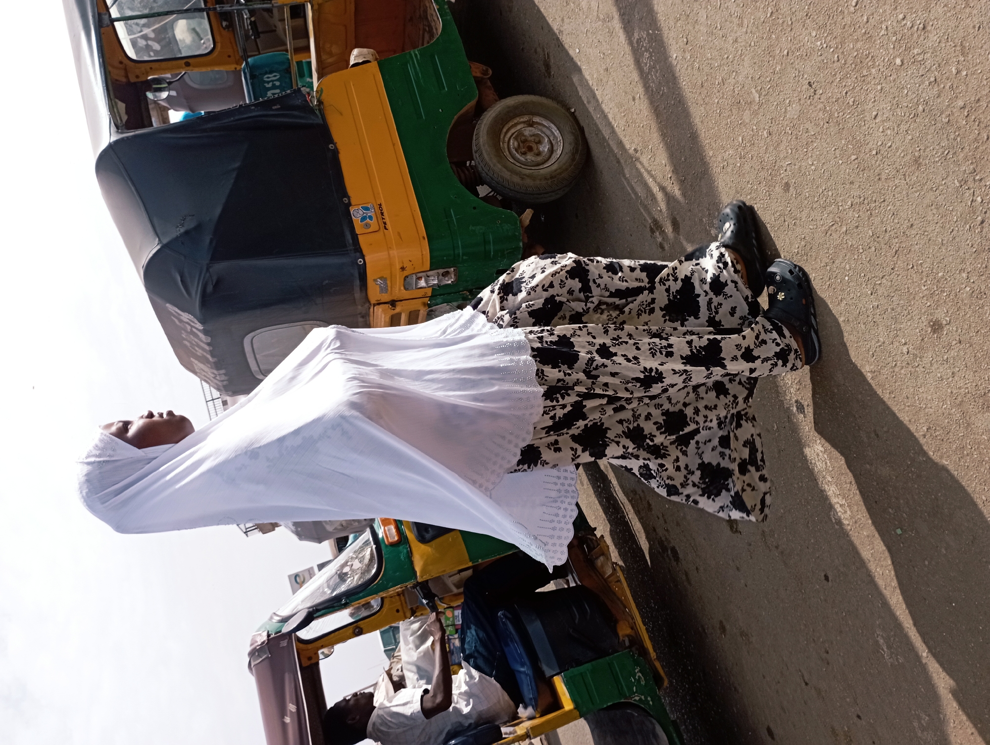 The image captures a vibrant street scene in Jos, Nigeria, featuring two people and auto-rickshaws. The main focus is a person standing beside a yellow and green auto-rickshaw, often called a 'keke napep'. This person is clad in a long, flowing white head covering or shawl, paired with a long, black and white floral patterned dress. Dark-colored clogs with small decorative charms adorn their feet. Their gaze is directed upwards, suggesting they might be looking at the sky or something in the distance. Distinct shadows on the light-colored, textured ground indicate it is daytime, likely a bright morning or afternoon.
Inside the auto-rickshaw, another individual, possibly the driver or a passenger, is visible. This person is wearing a white shirt and appears to be looking down, perhaps at a phone or object in their hands. The auto-rickshaw itself is a common three-wheeled vehicle, with a black fabric covering over the passenger compartment. A blue sticker on its yellow body reads "PETROL", and another blue container inside the vehicle shows the numbers "850". A portion of a second auto-rickshaw is also visible in the lower left, further establishing the setting as a bustling street. The sky above is uniformly bright white, hinting at a clear or hazy day. The overall impression is a snapshot of everyday life and transport in an urban environment in Jos.