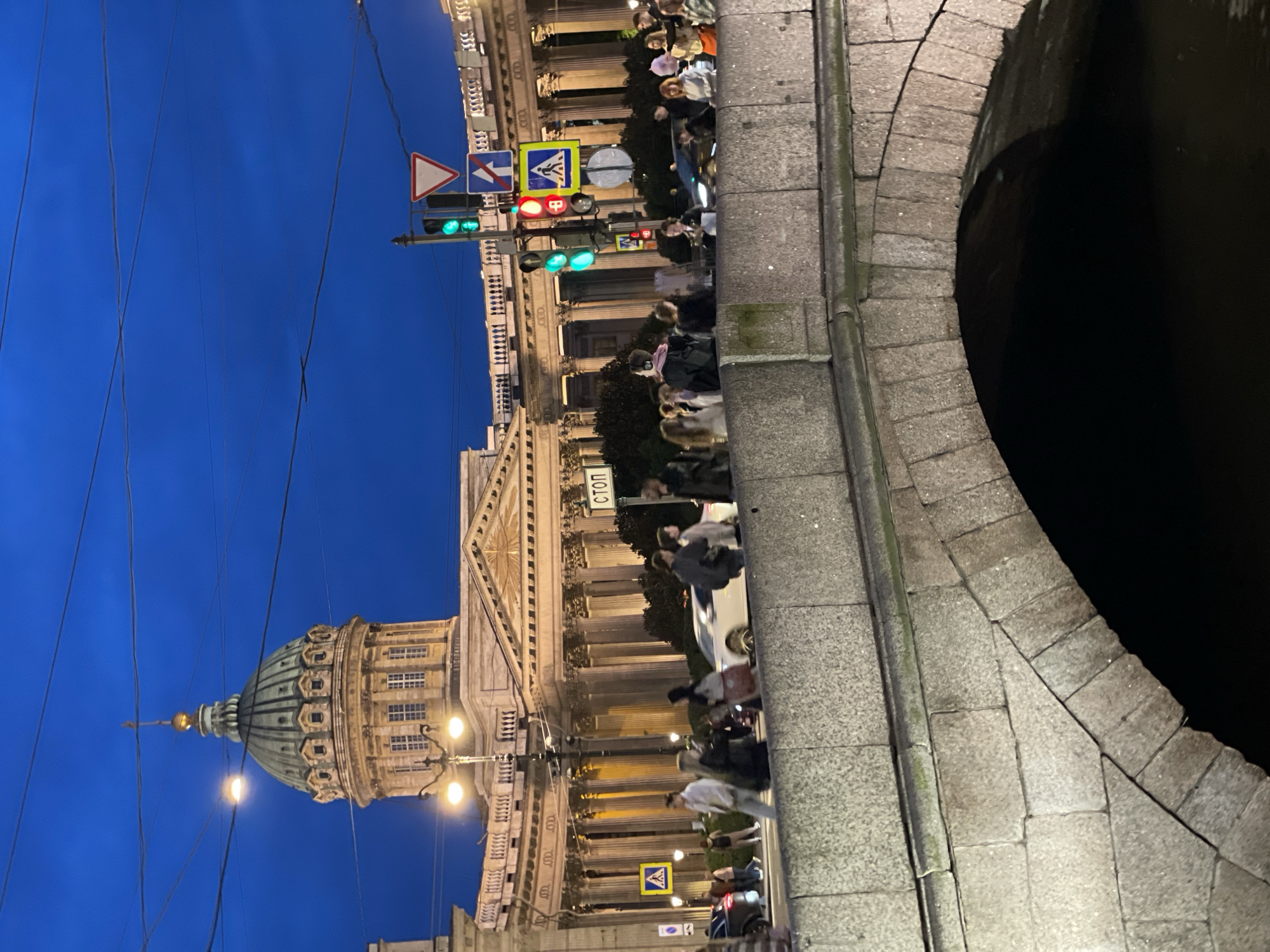 The image depicts a night scene in front of Kazan Cathedral in St. Petersburg, Russia. A stone bridge spans a dark body of water in the foreground, with its arch visible. Behind the bridge, the grand facade of Kazan Cathedral, characterized by its colonnade and dome, is illuminated. A street with traffic lights and pedestrian crossing signs is visible, with several people walking and cars passing by. The sky is a deep blue, indicating twilight or night.