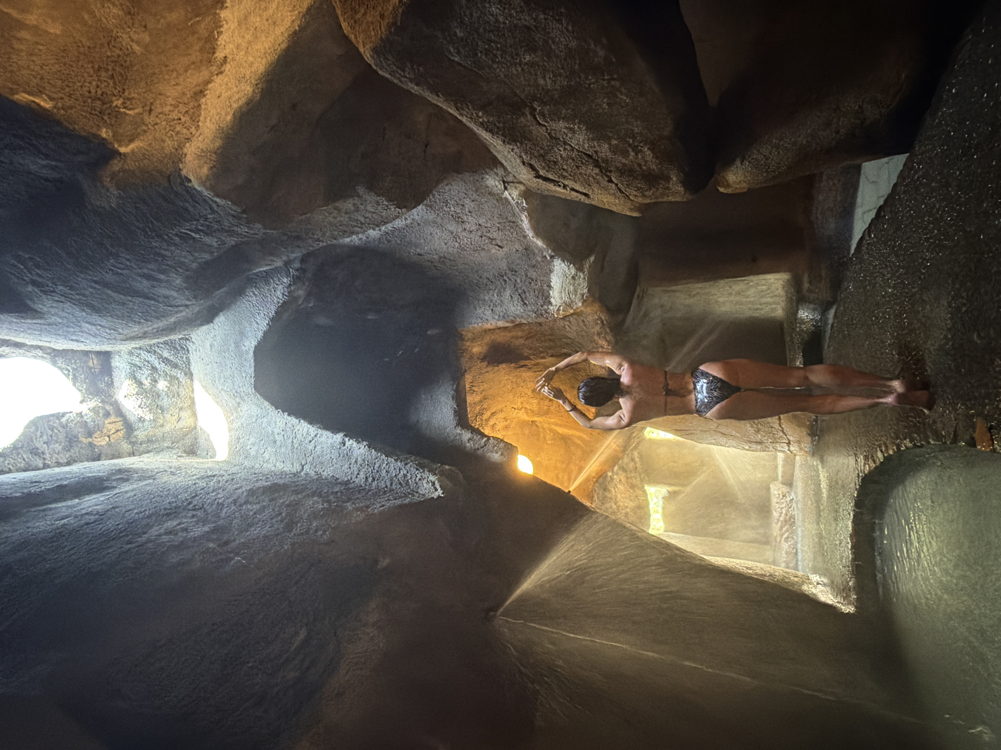 The image shows a woman from behind, wearing a patterned bikini. She is standing in what appears to be a natural cave or grotto, with rough, textured rock formations forming the walls and ceiling. Sunlight streams in from openings in the ceiling, illuminating the space with a warm, golden light. Water is visible on the ground, suggesting a humid environment, and a mist or spray is present in the air, particularly to the left of the woman. She has her arms raised above her head, with her hands clasped together, in a pose that could be stretching or relaxing. The overall impression is one of a serene and natural spa-like setting. The location is identified as being in the city of Nha Trang Ward, Vietnam.